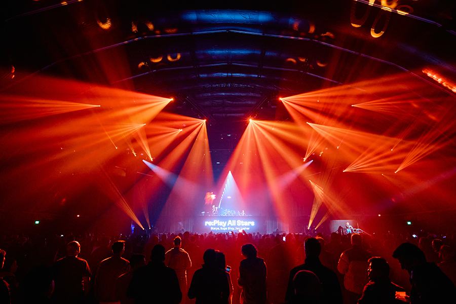 A crowd of people standing around a stage with red and yellow lights.