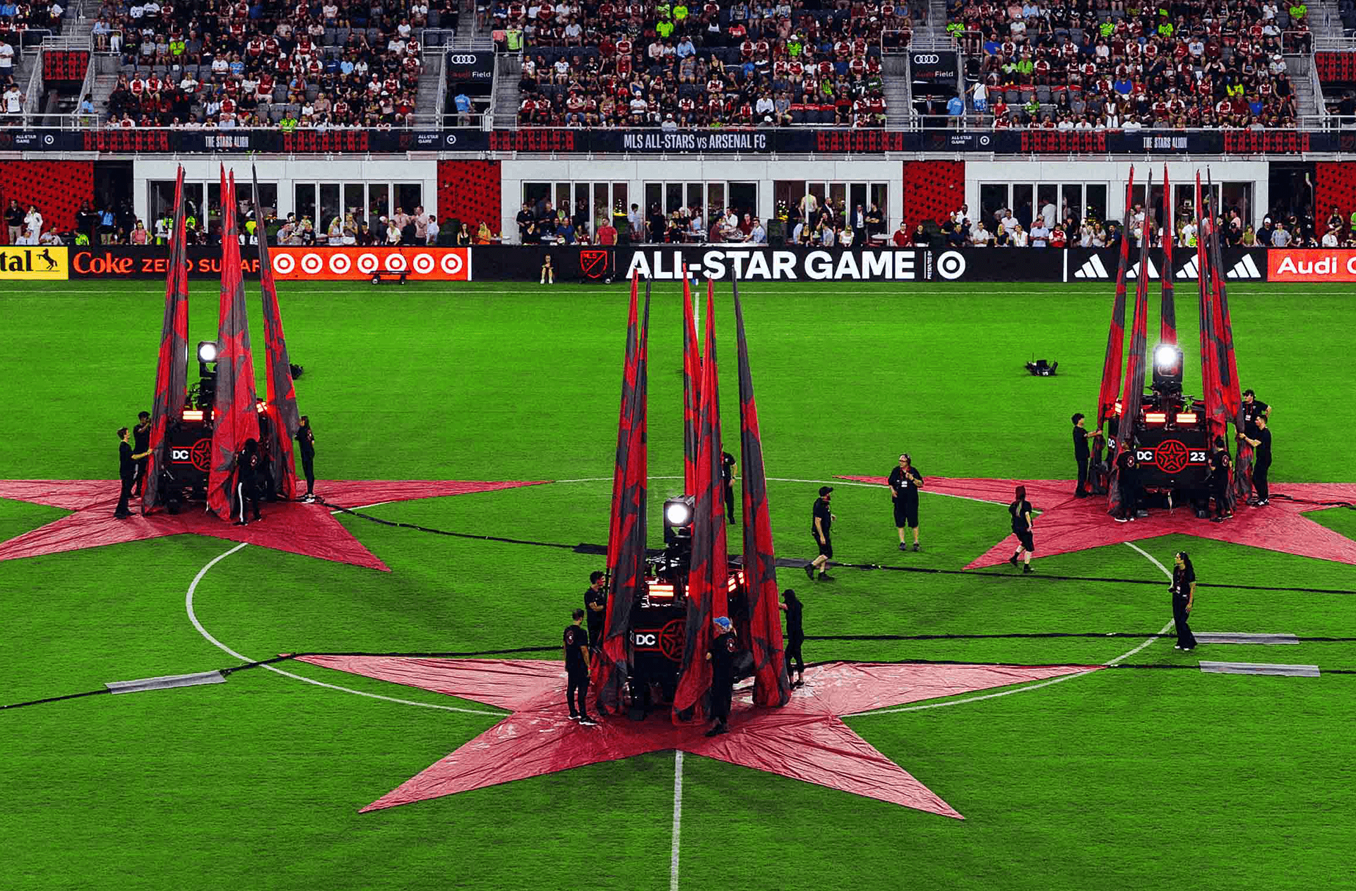 MLS All-Star Game opening ceremony with three large red stars and towers on a green soccer field surrounded by a crowd.