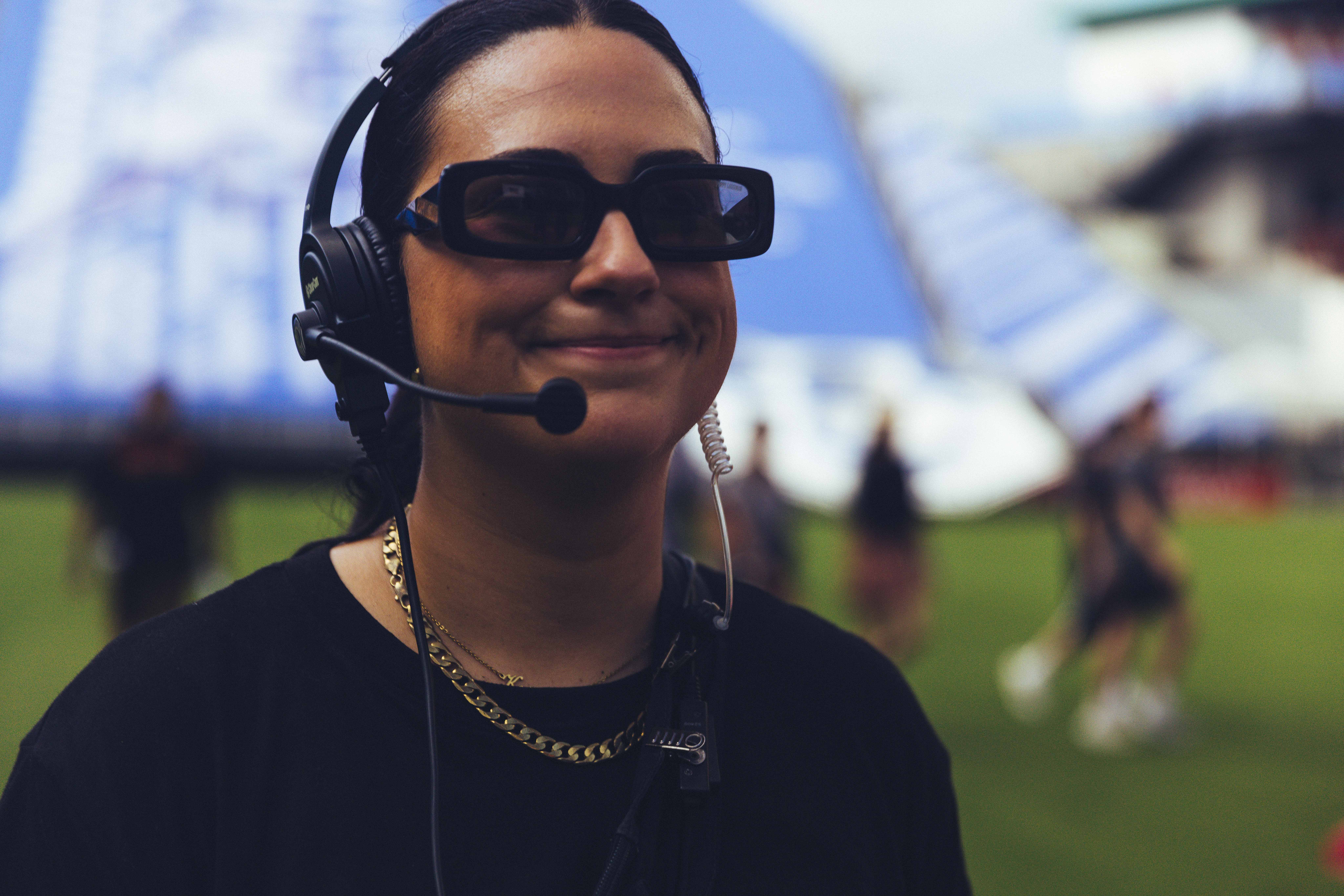 a woman dressed in a black t-shirt with sunglasses and a headset with grass in the background