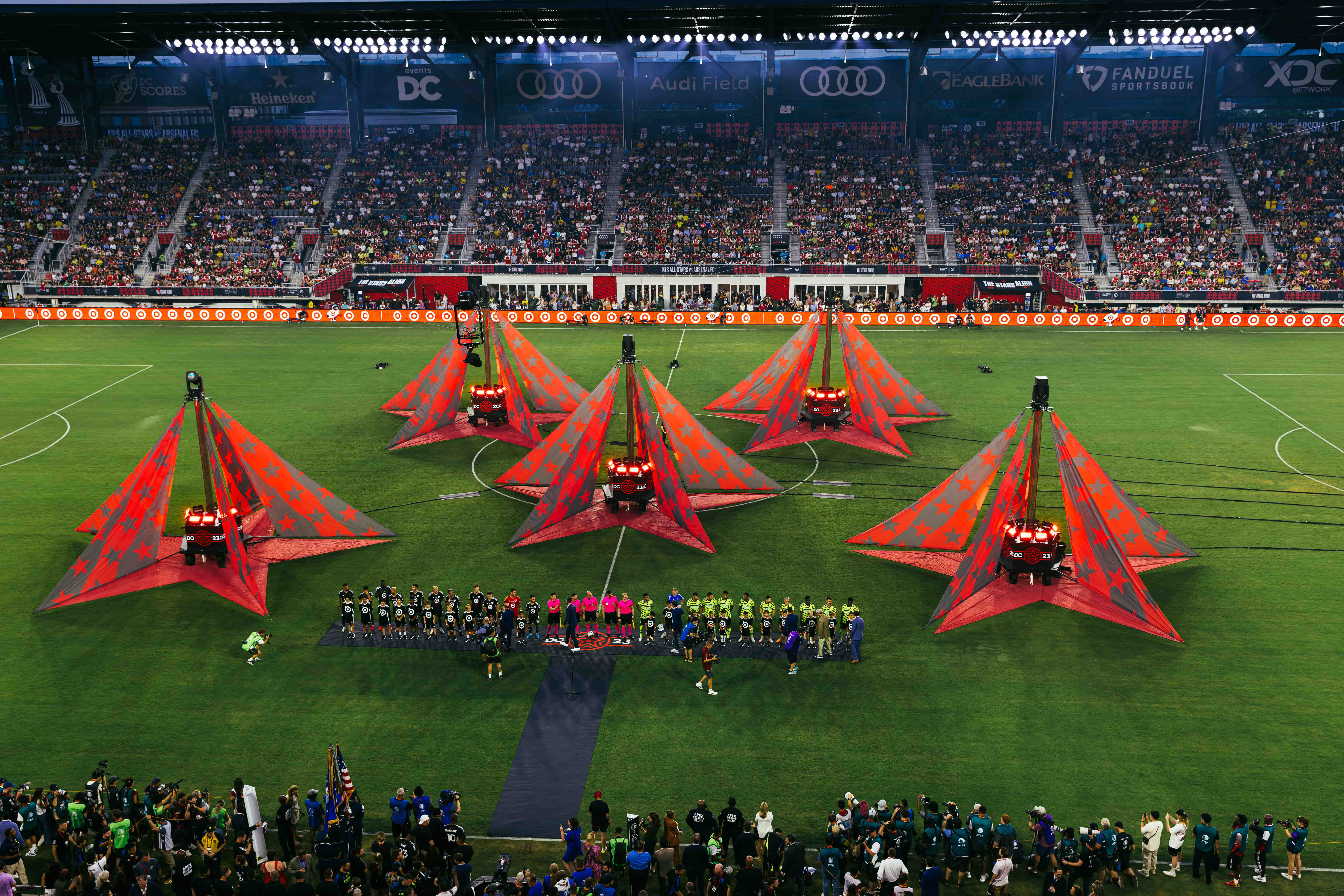 Two soccer teams lined up on a field with large red star-shaped decorations and a crowd in stadium stands at Audi Field.