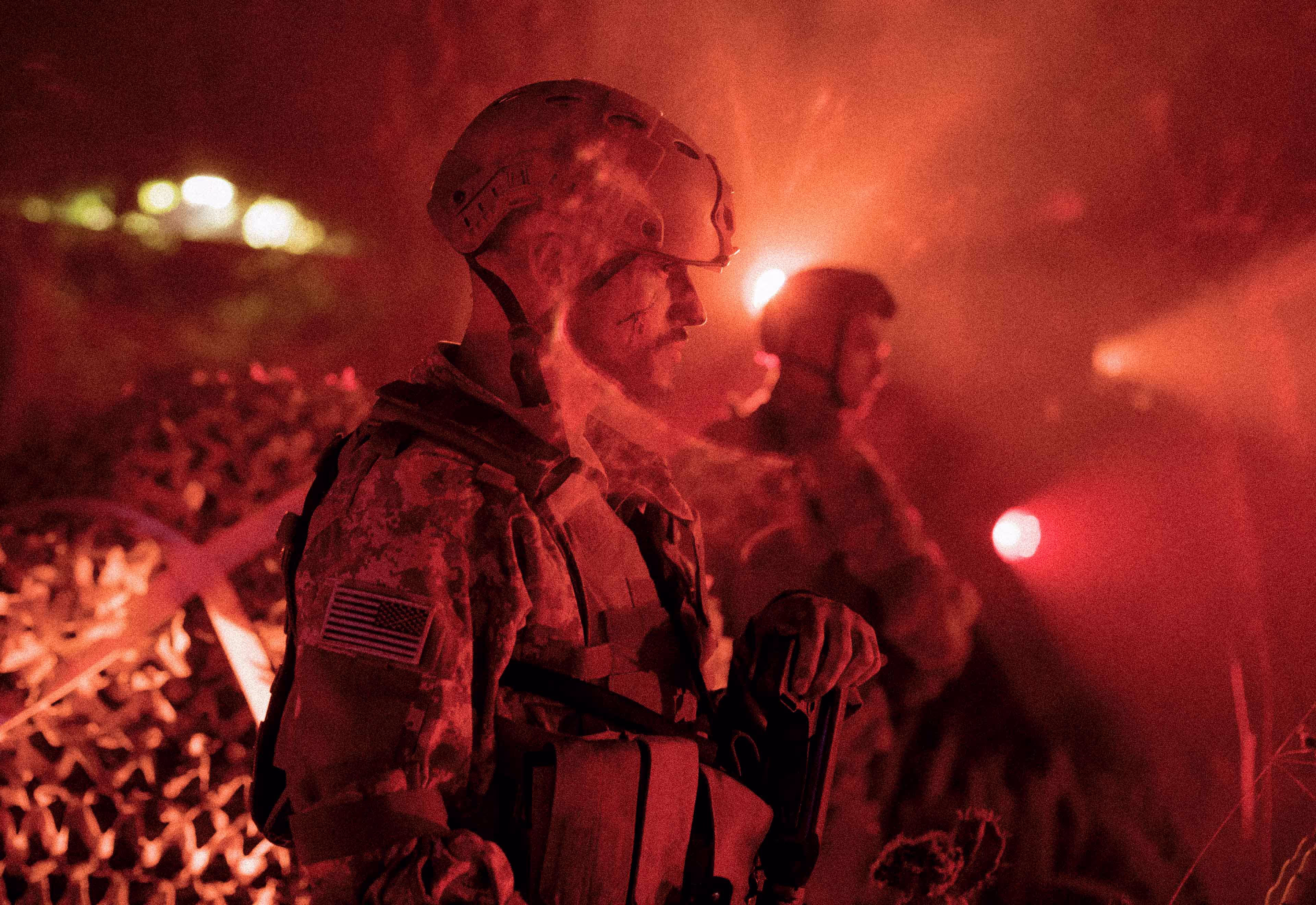 Soldiers in tactical gear and helmets standing with red light and smoke in the background.