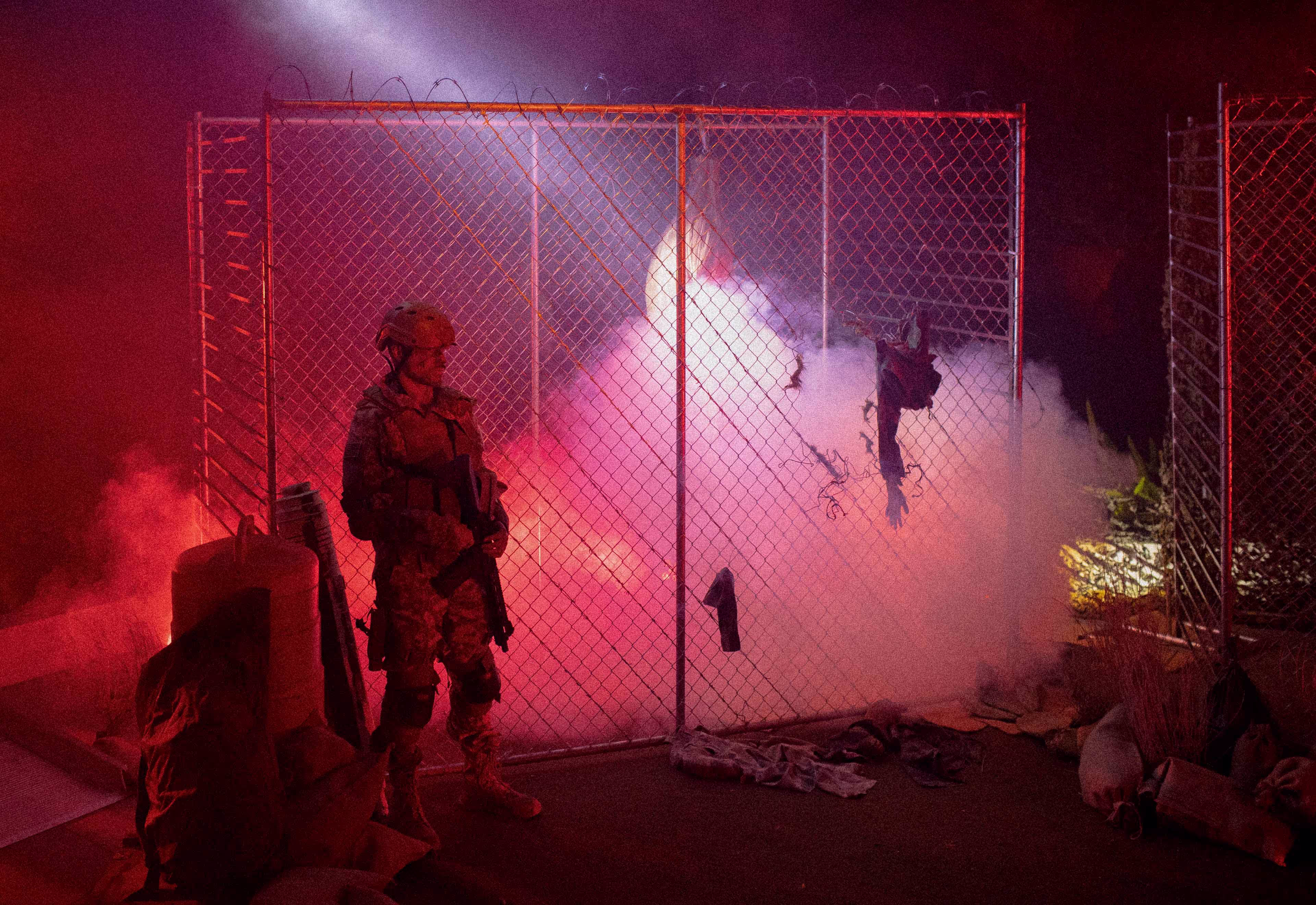 Soldier in camouflage and helmet standing guard beside a barbed wire fence with smoke and red lighting in the background at night.