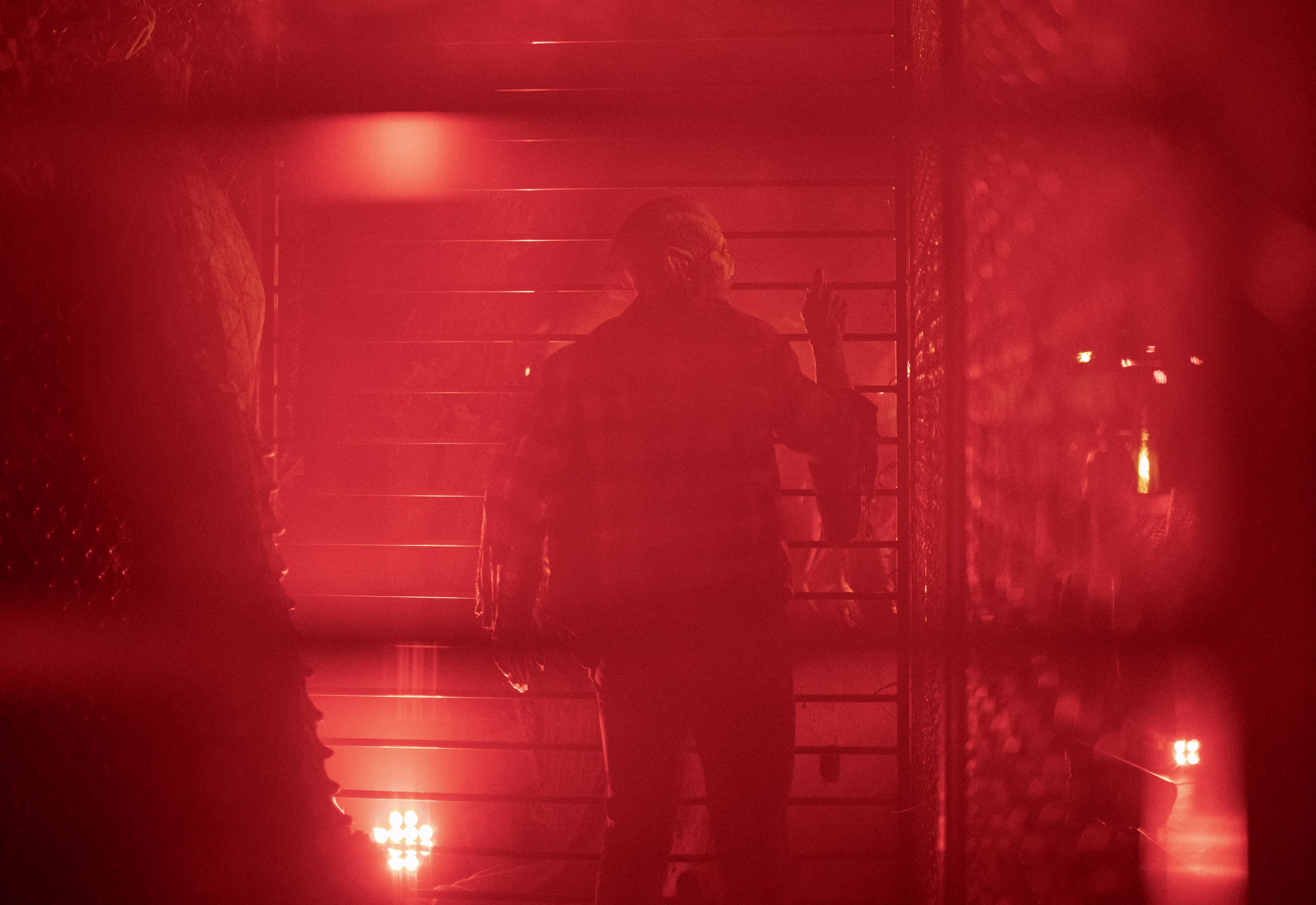 Silhouette of a person with pointed ears standing behind a metal fence, surrounded by red lighting and smoke.