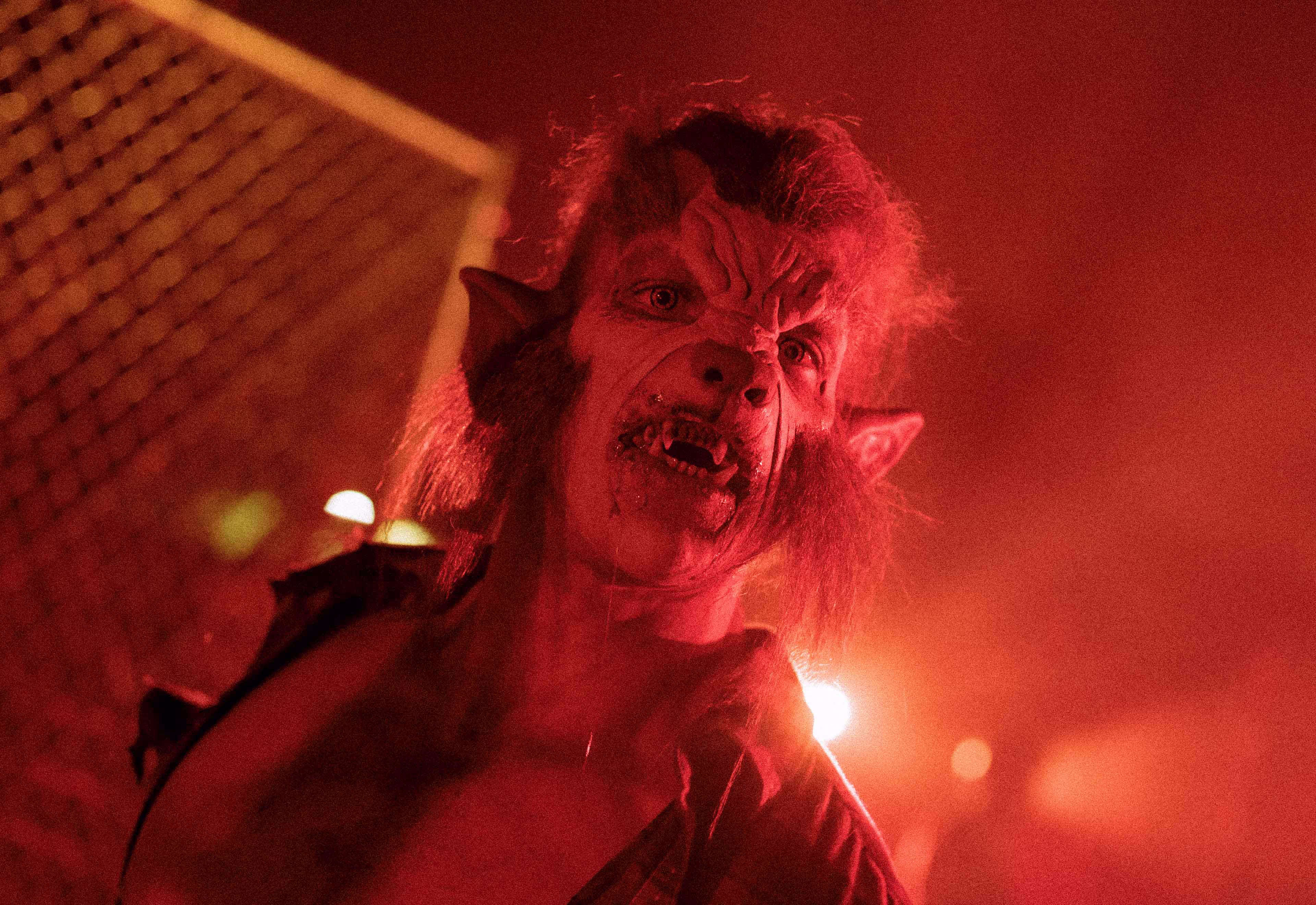 Person in a detailed werewolf costume with fangs and pointed ears under red lighting.