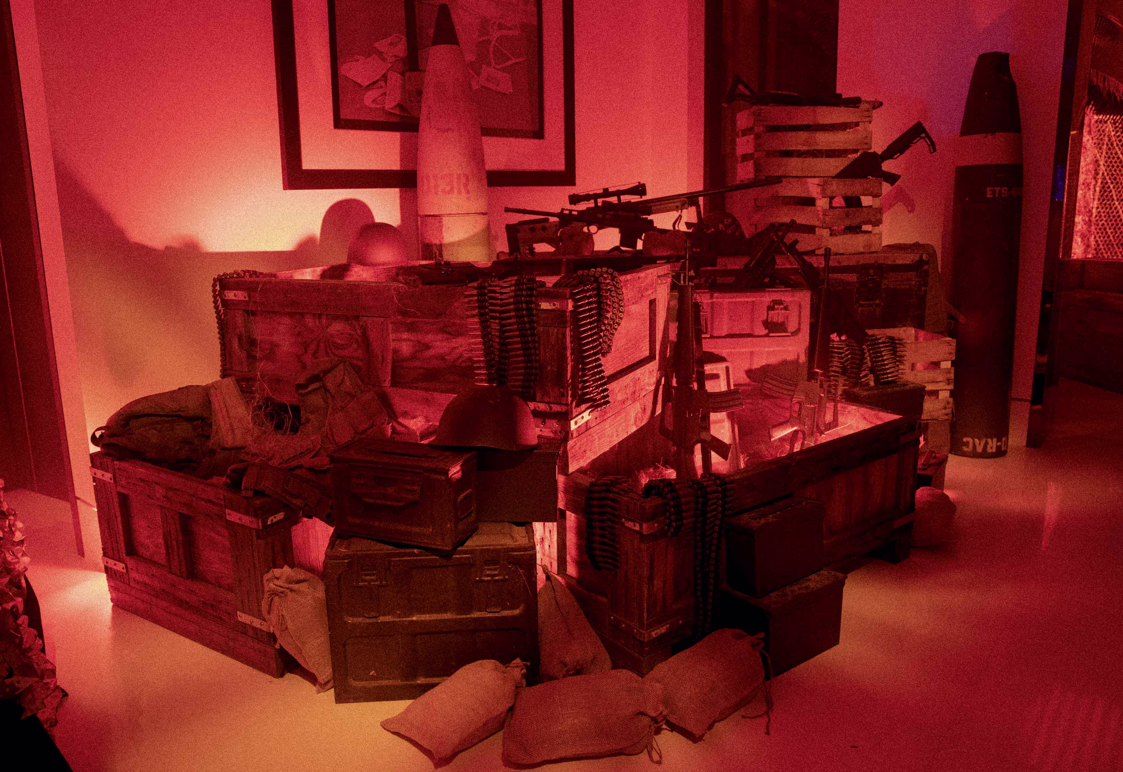 Dimly lit room with military crates, helmets, rifles, ammunition belts, sandbags, and large artillery shells.