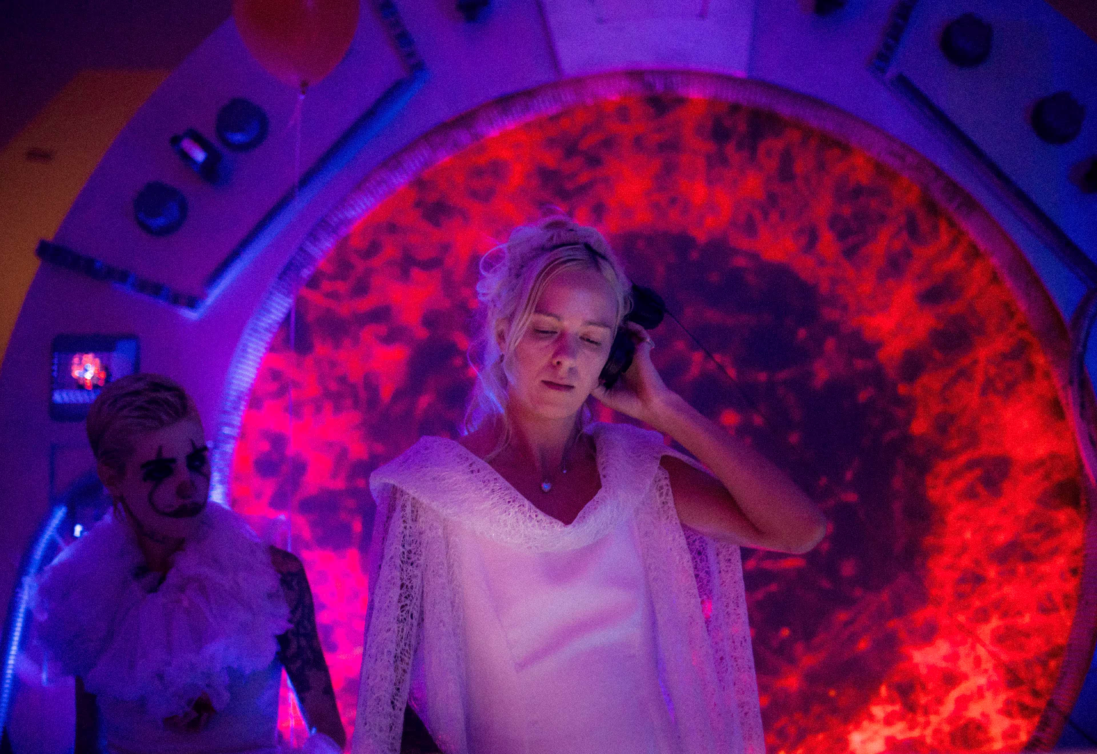 A woman in a white dress and lace cape wearing headphones in front of a glowing red circular background.
