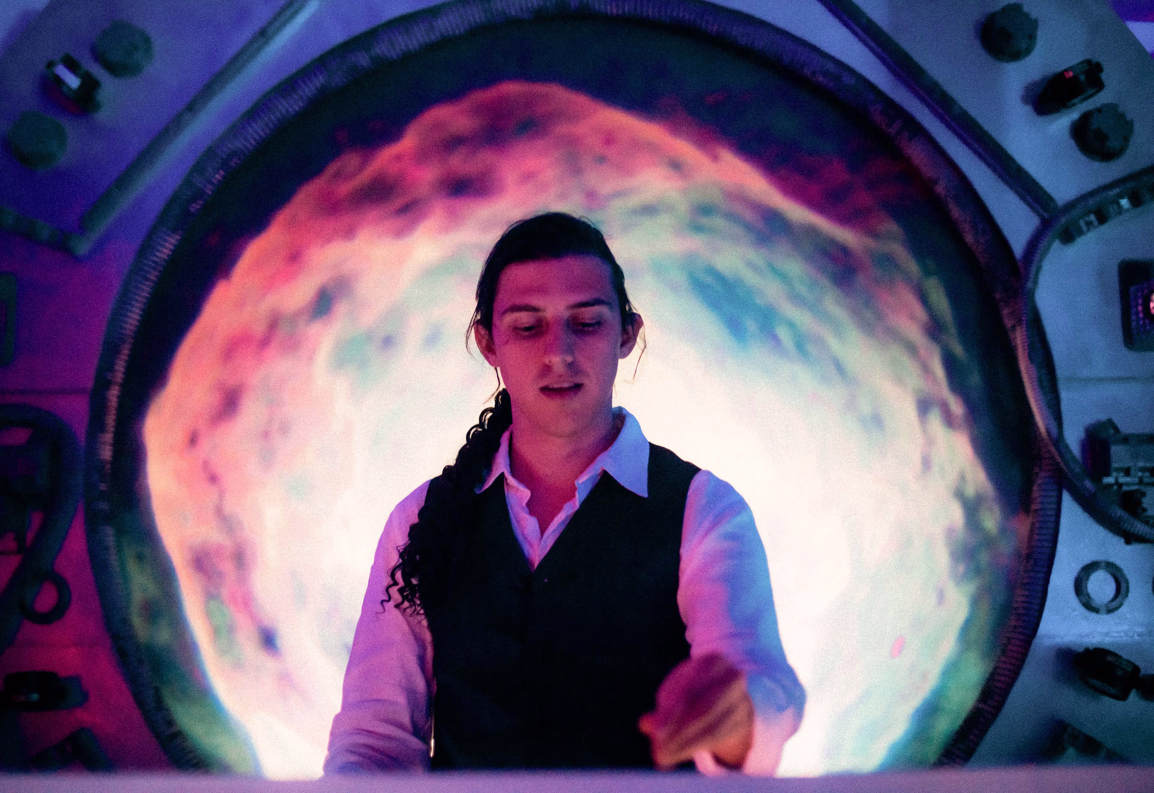 Man with long curly hair tied back in front of a circular colorful light display.