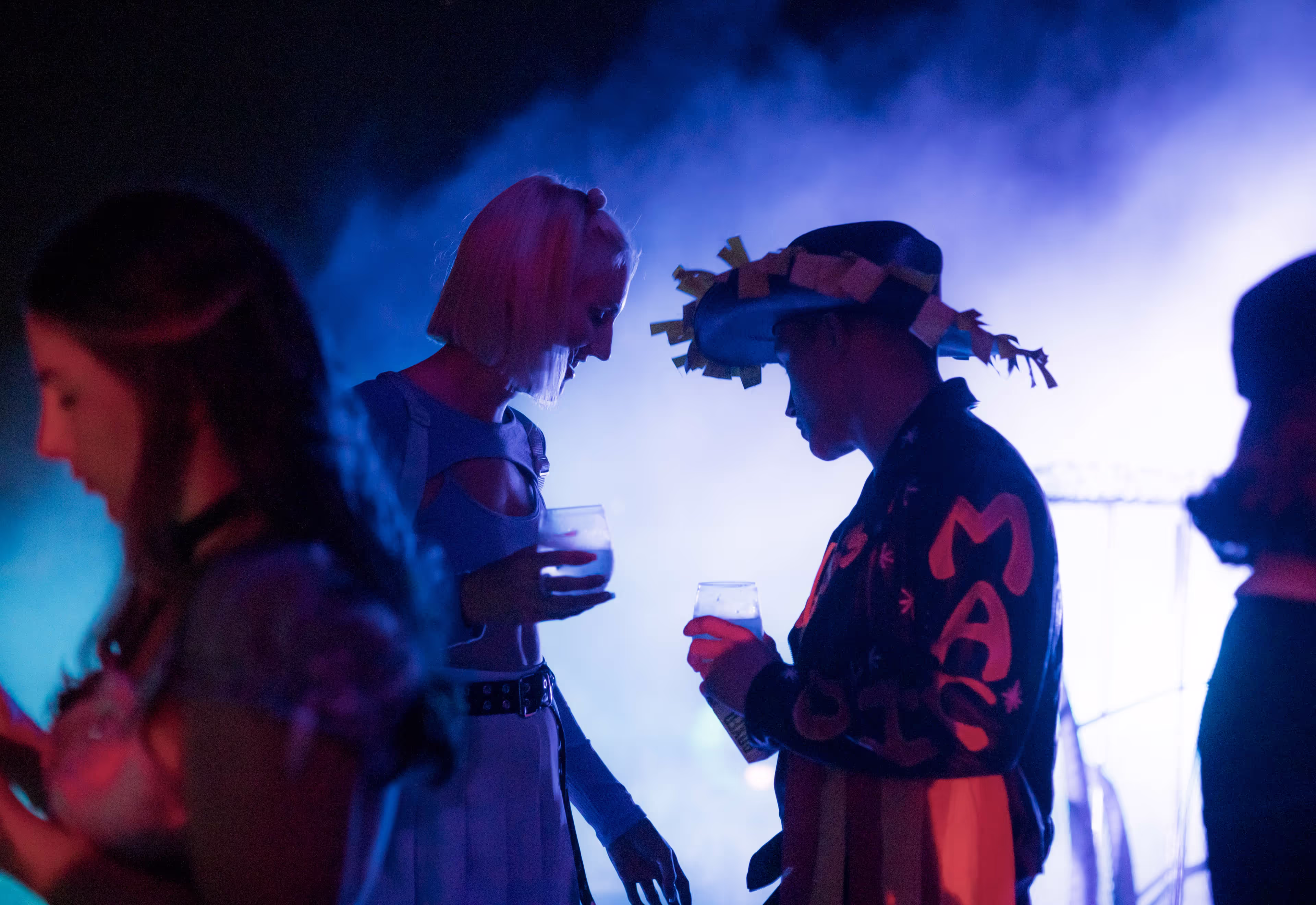Two people in costumes sharing drinks at a party with blue and purple lighting.