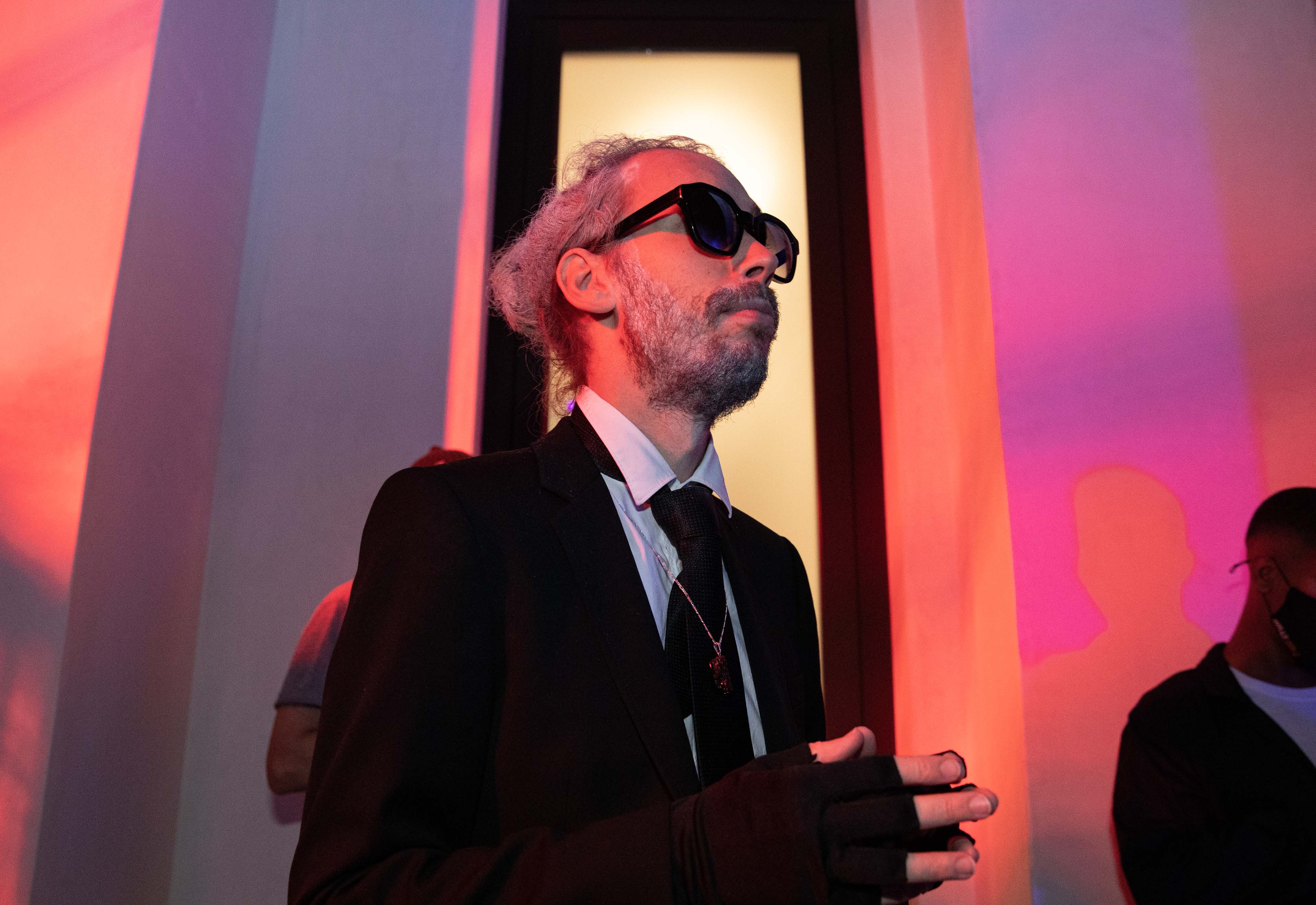 Man with gray hair in a bun and beard wearing black sunglasses and a black suit.