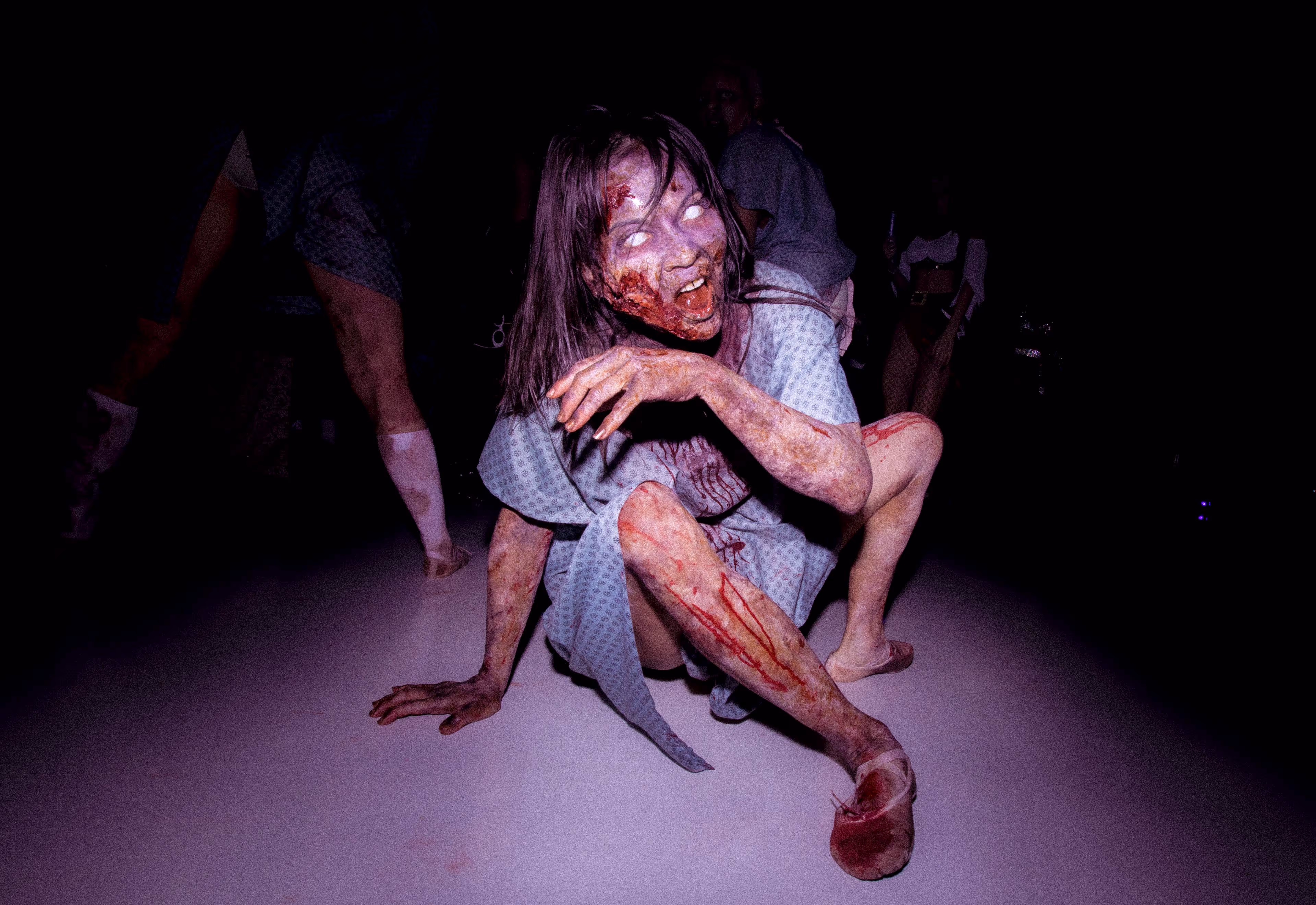 Person in a bloodied hospital gown crouching on the floor with pale zombie-like makeup.