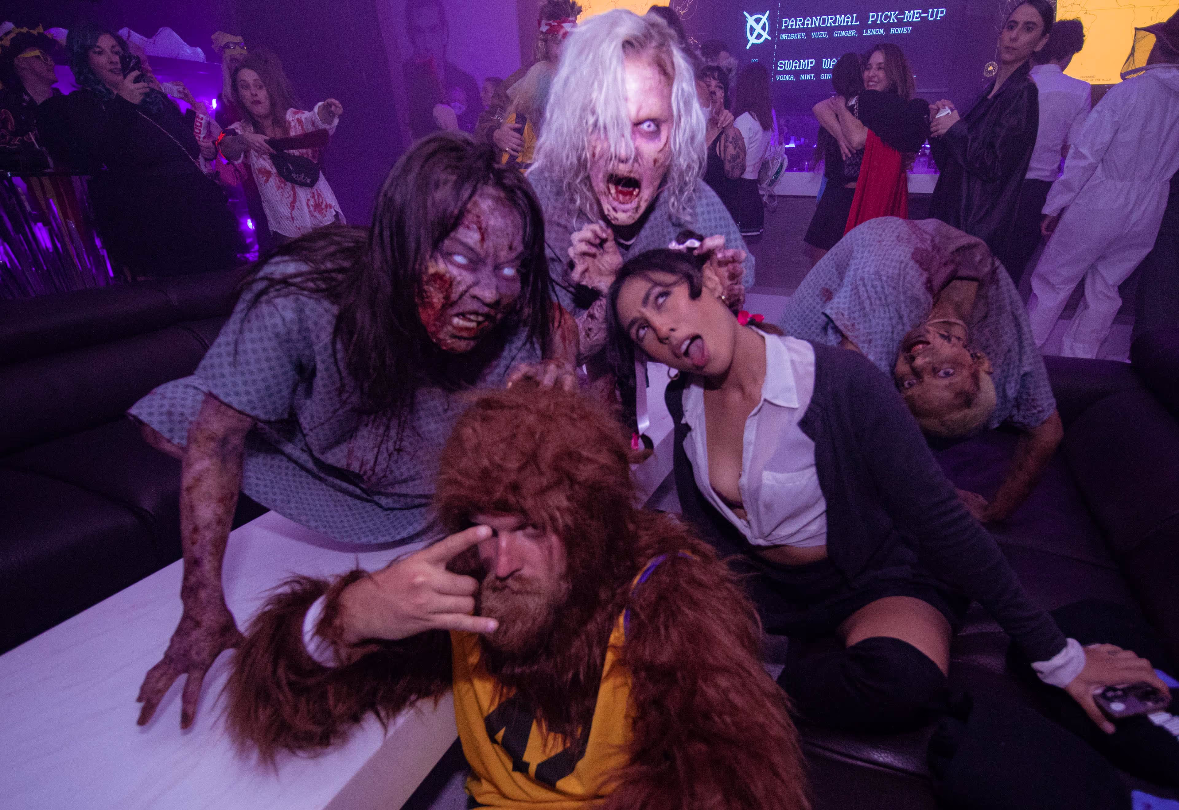 Group of people in Halloween costumes including zombies and a creature with brown fur.
