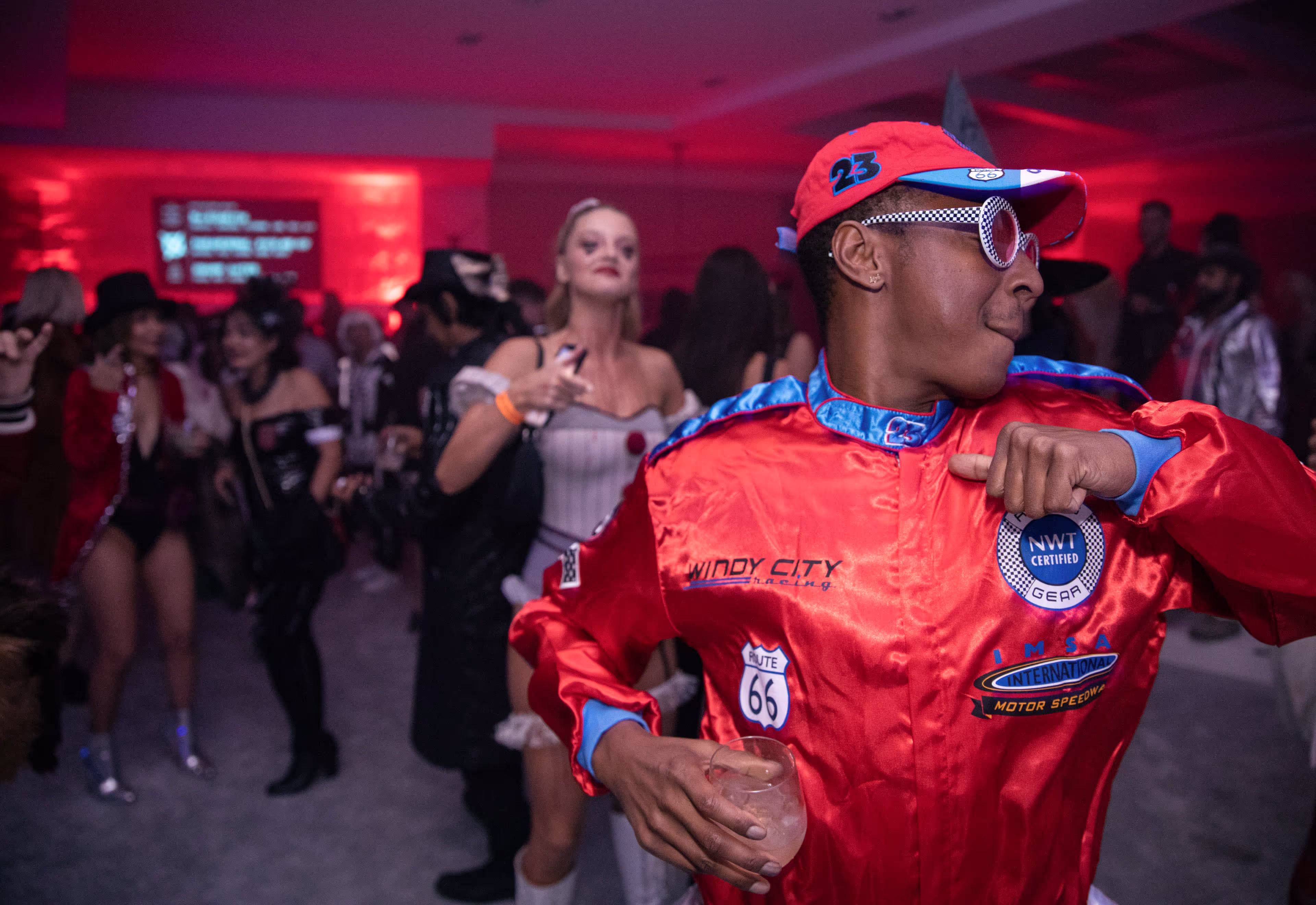 Person in a red racing jacket and cap dancing at a party with colorful lights.