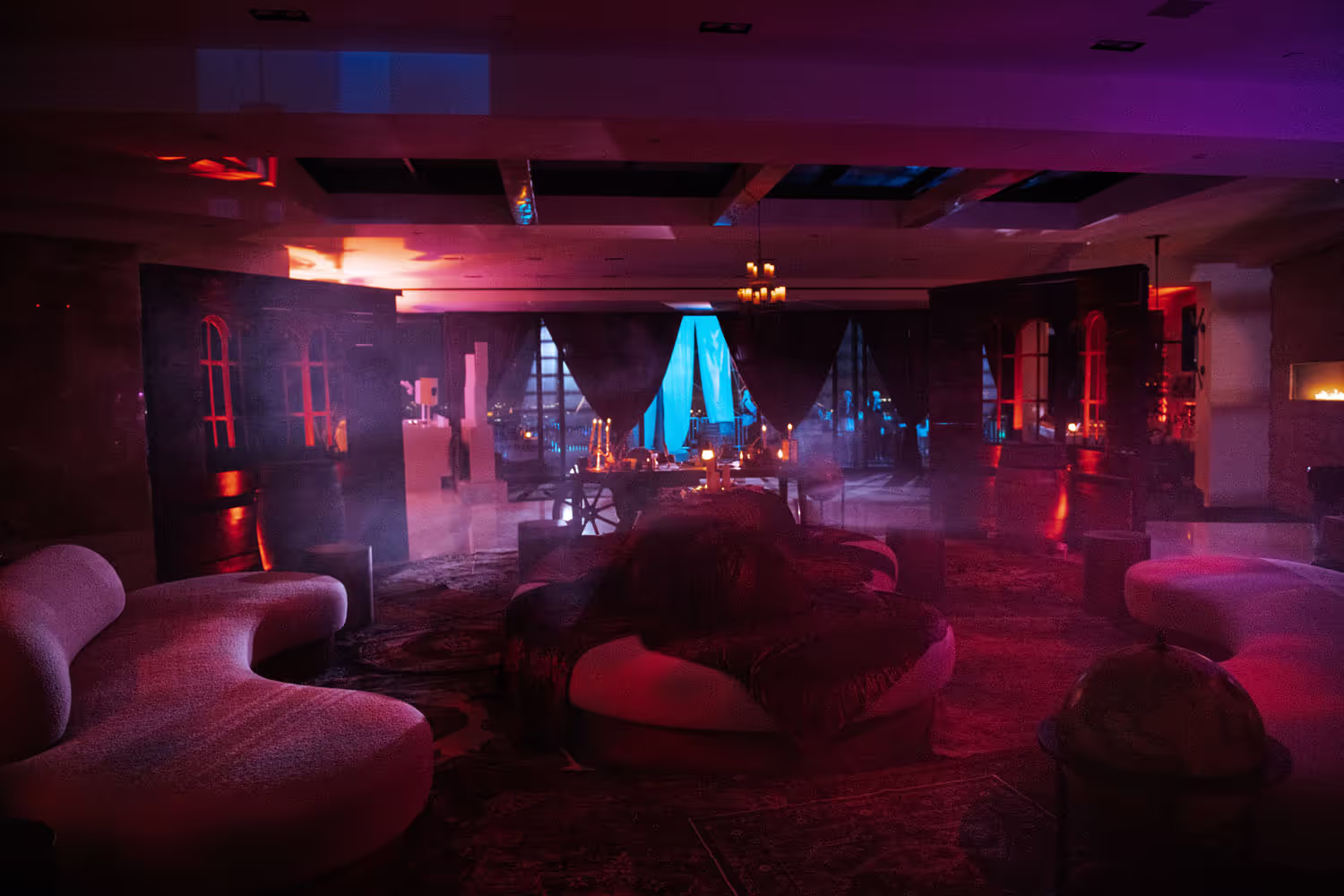 Ambient dimly lit room with curved sofas, central round bed, candles, and large windows with dark curtains.