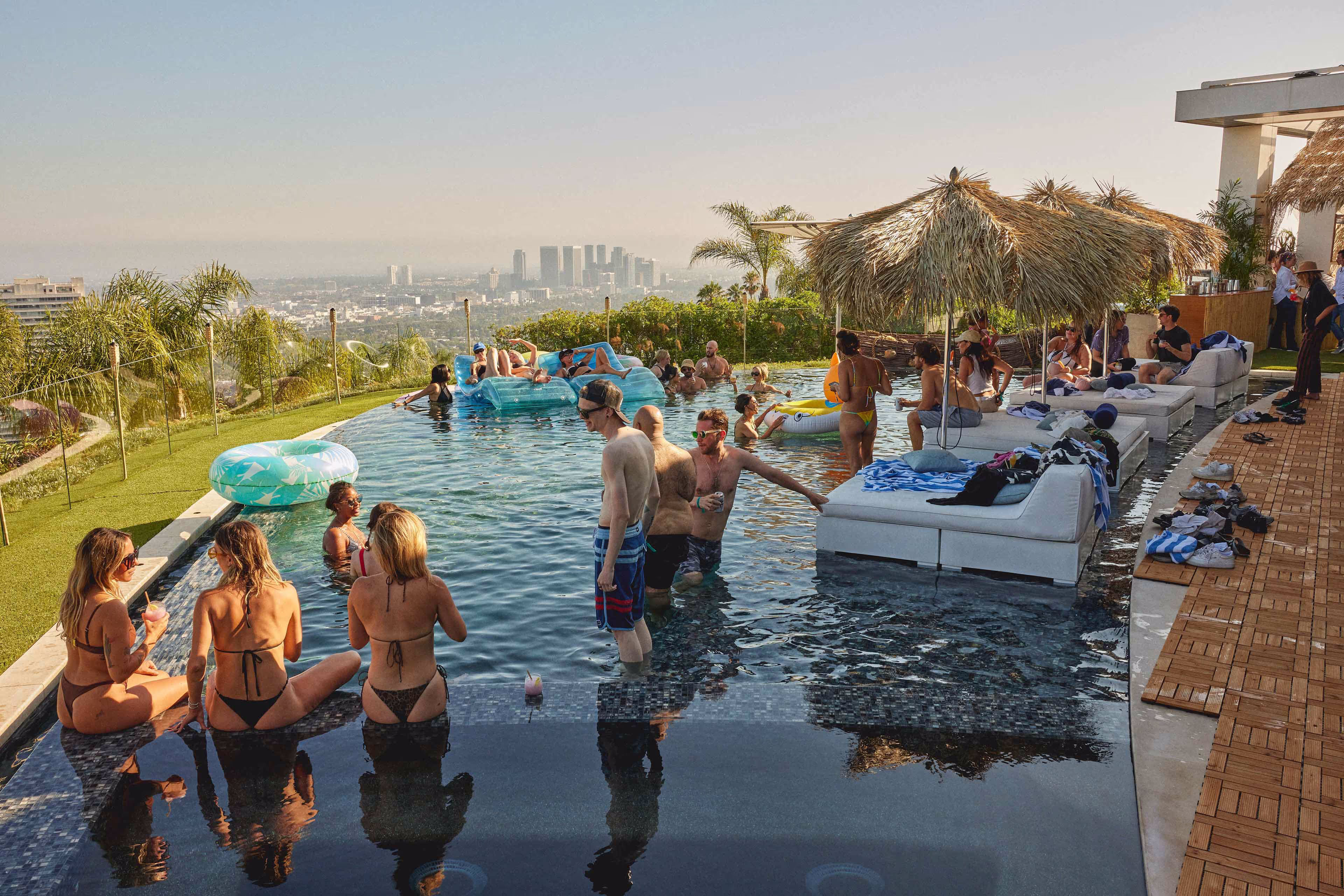 People socializing and relaxing in an infinity pool with city skyline and palm trees in the background.
