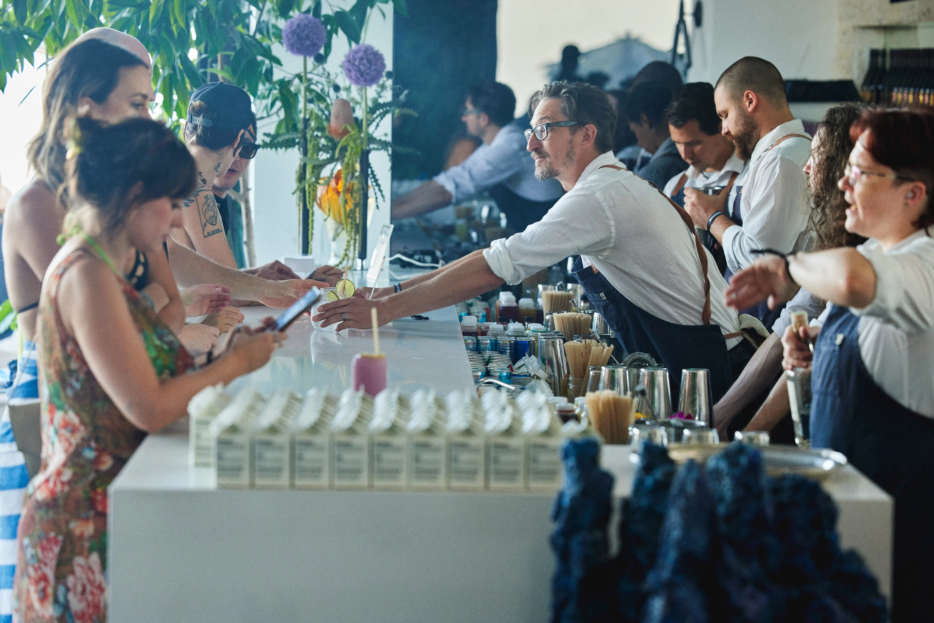Bartenders wearing aprons serve drinks to customers seated along a white bar counter decorated with plants and drink ingredients.
