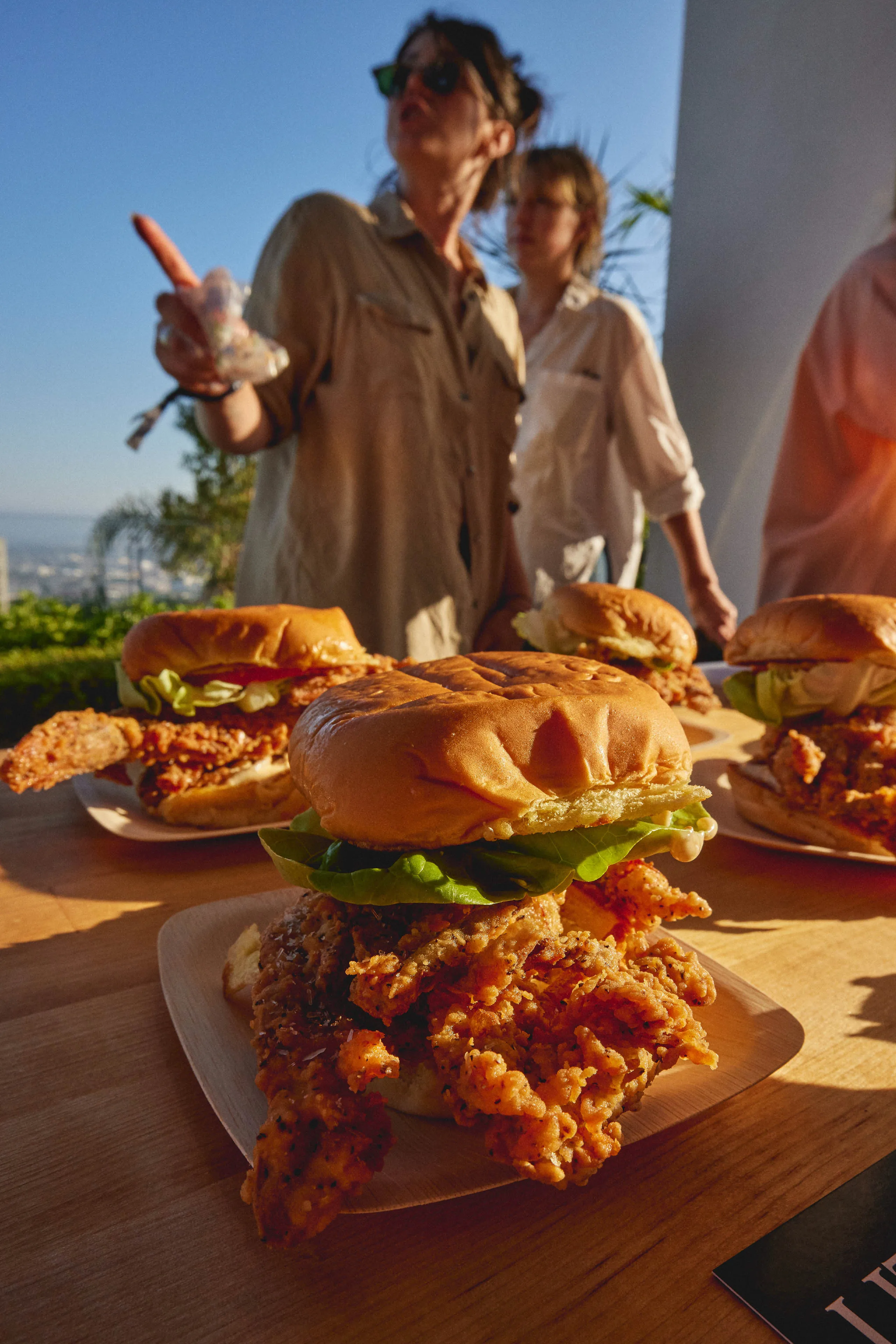 Close-up of crispy fried chicken sandwich with lettuce on a wooden plate, with people and more sandwiches in the background.