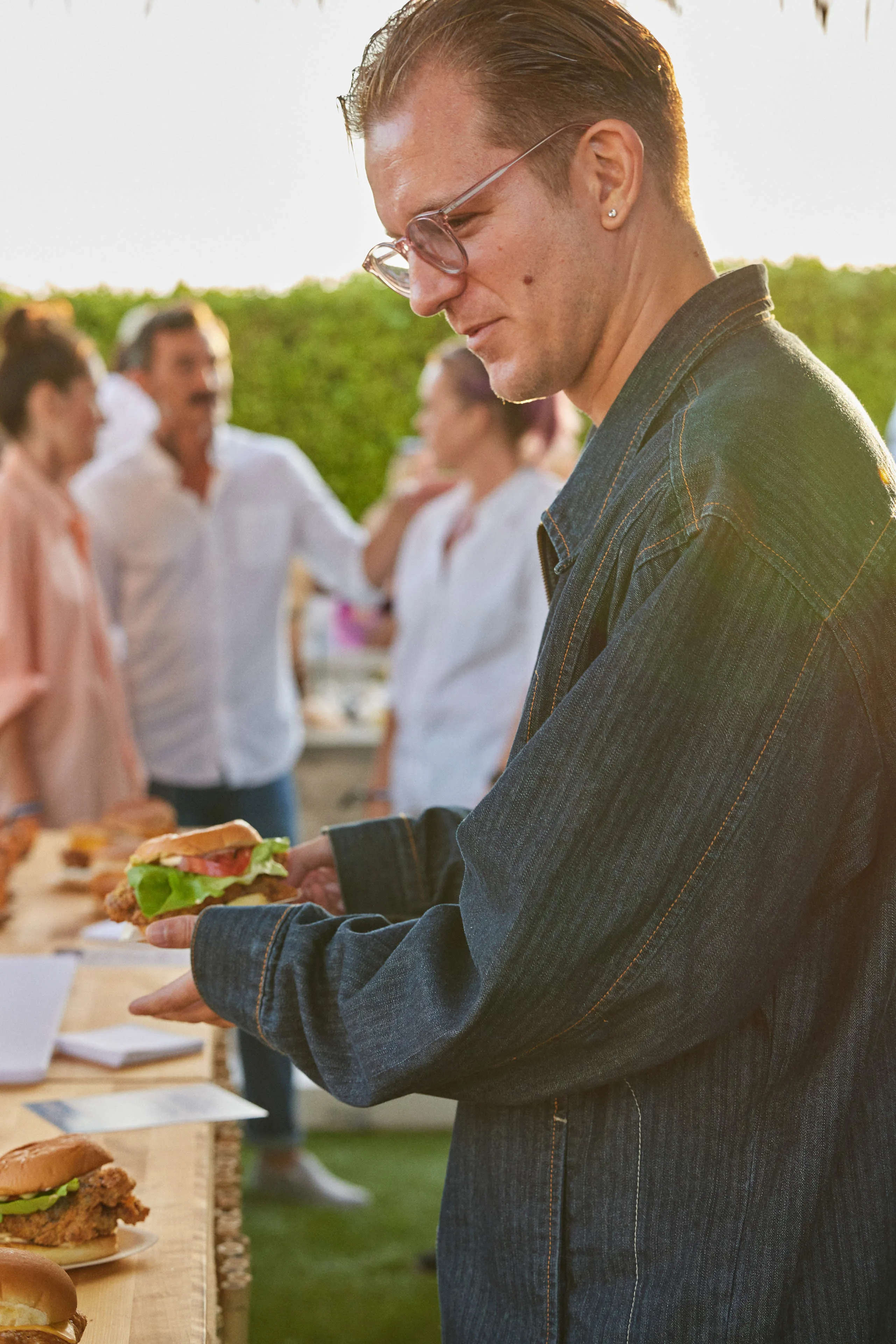 Man wearing glasses and a denim jacket holding a chicken sandwich at an outdoor gathering with other people in the background.