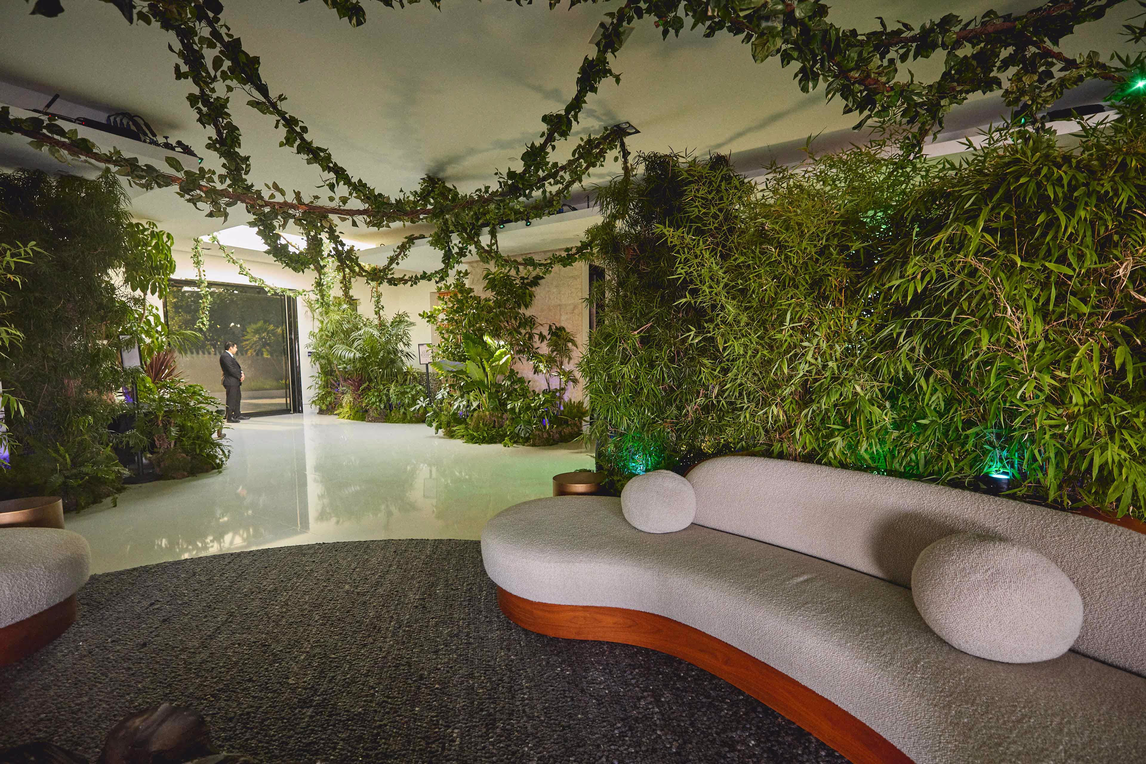 Curved modern sofa with round cushions beside a lush green wall and hanging vines in a spacious indoor area with reflective flooring.