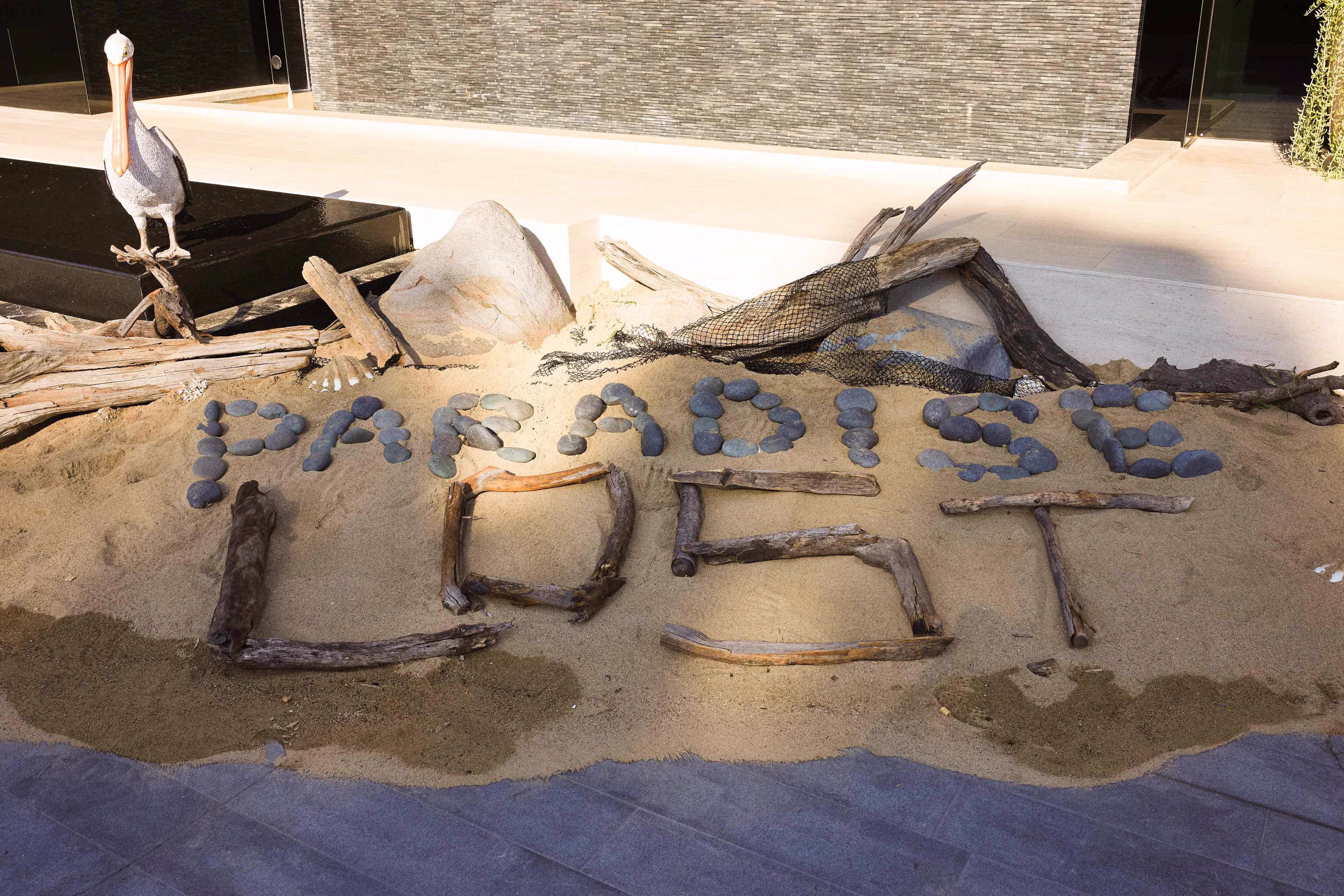 Arrangement of stones and sticks on sand spelling 'PARADISE LOST' with a pelican statue standing on driftwood nearby.
