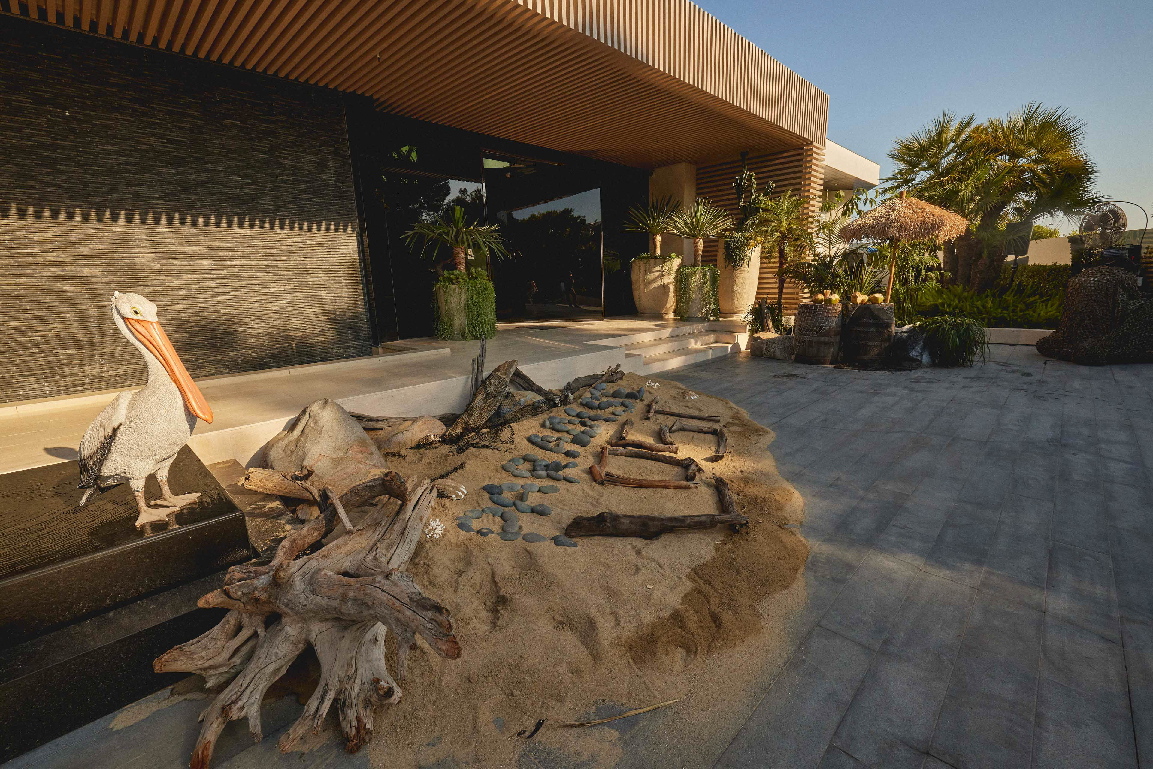 Outdoor modern patio with sand art featuring stones and driftwood spelling 'PARA LOST', potted plants, and a pelican sculpture on a black pedestal.