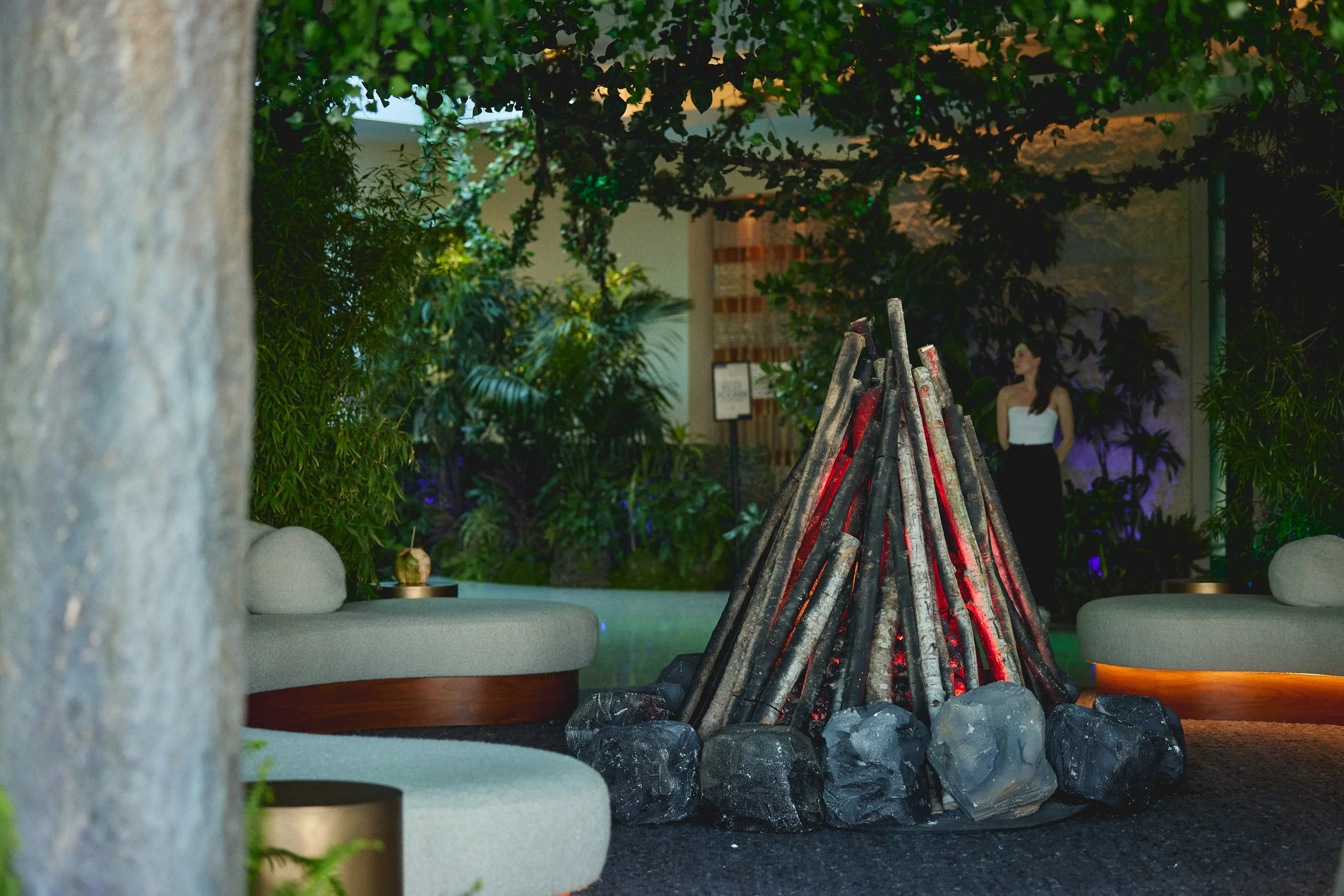 Modern indoor seating area with circular gray cushioned benches surrounding a large decorative fire pit made of vertical logs and black stones, with plants and a woman standing in the background.