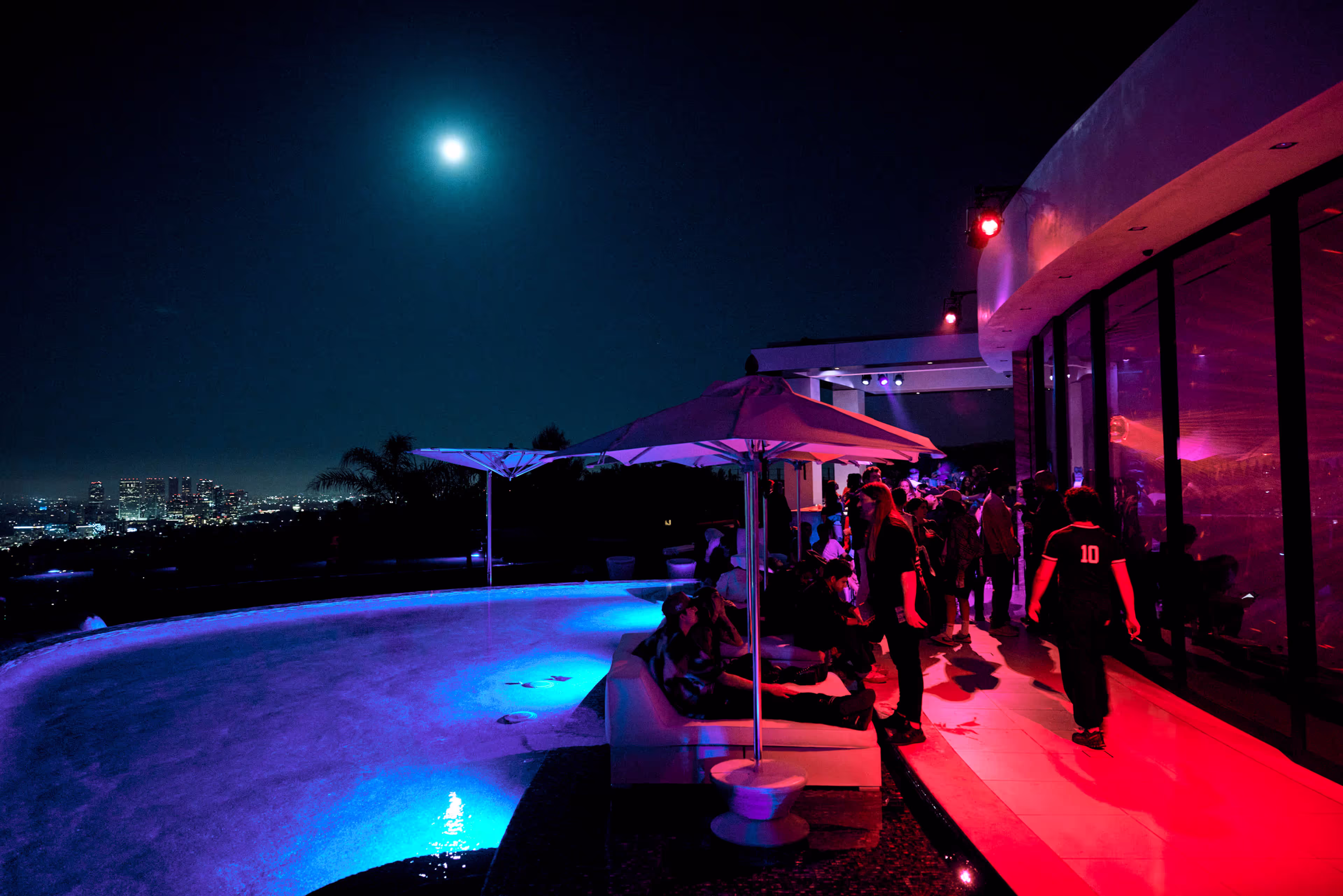 Nighttime rooftop party with people socializing near an illuminated pool and city skyline under a full moon.