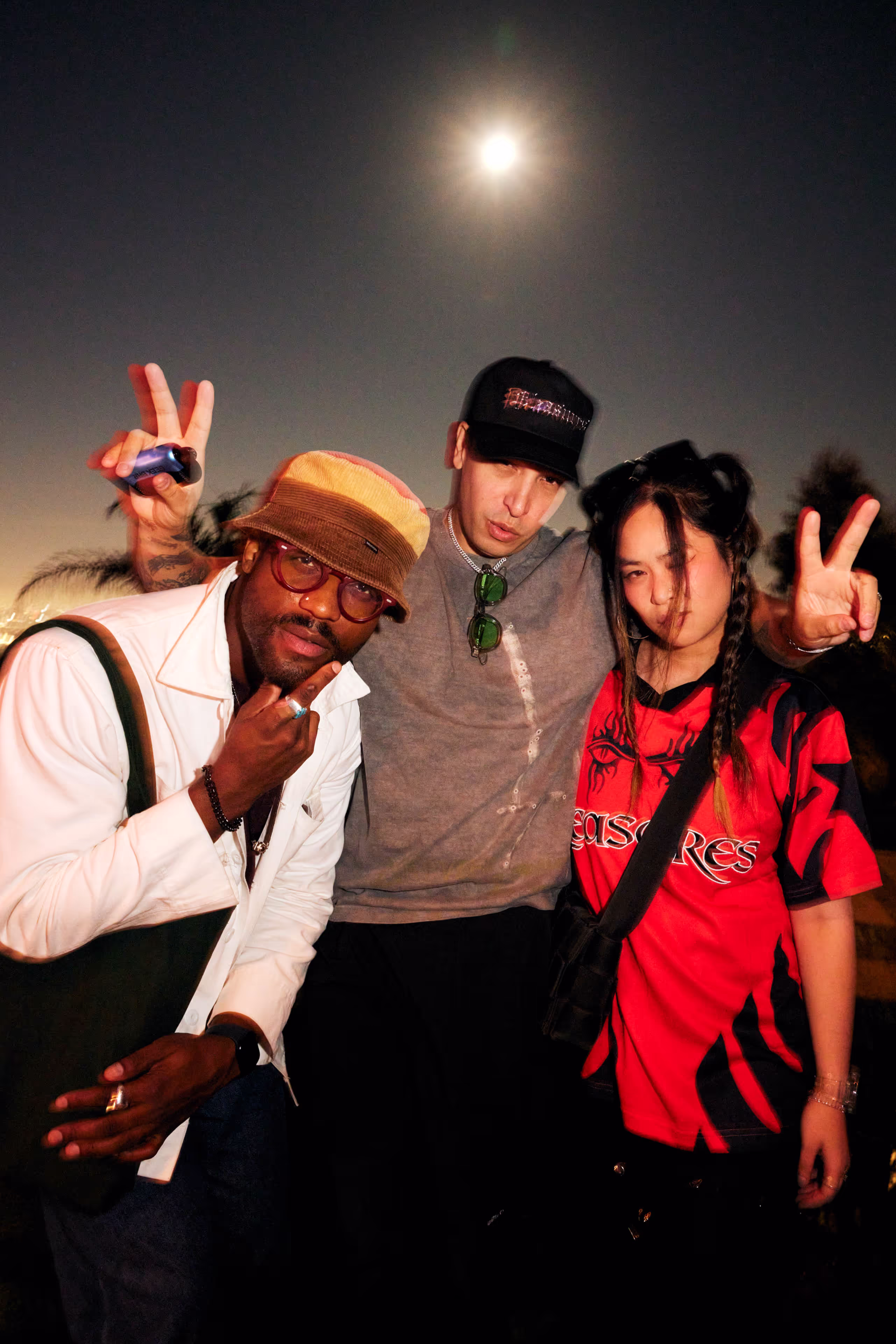 Three friends posing under the night sky with a bright moon, two making peace signs and one wearing a bucket hat and glasses.