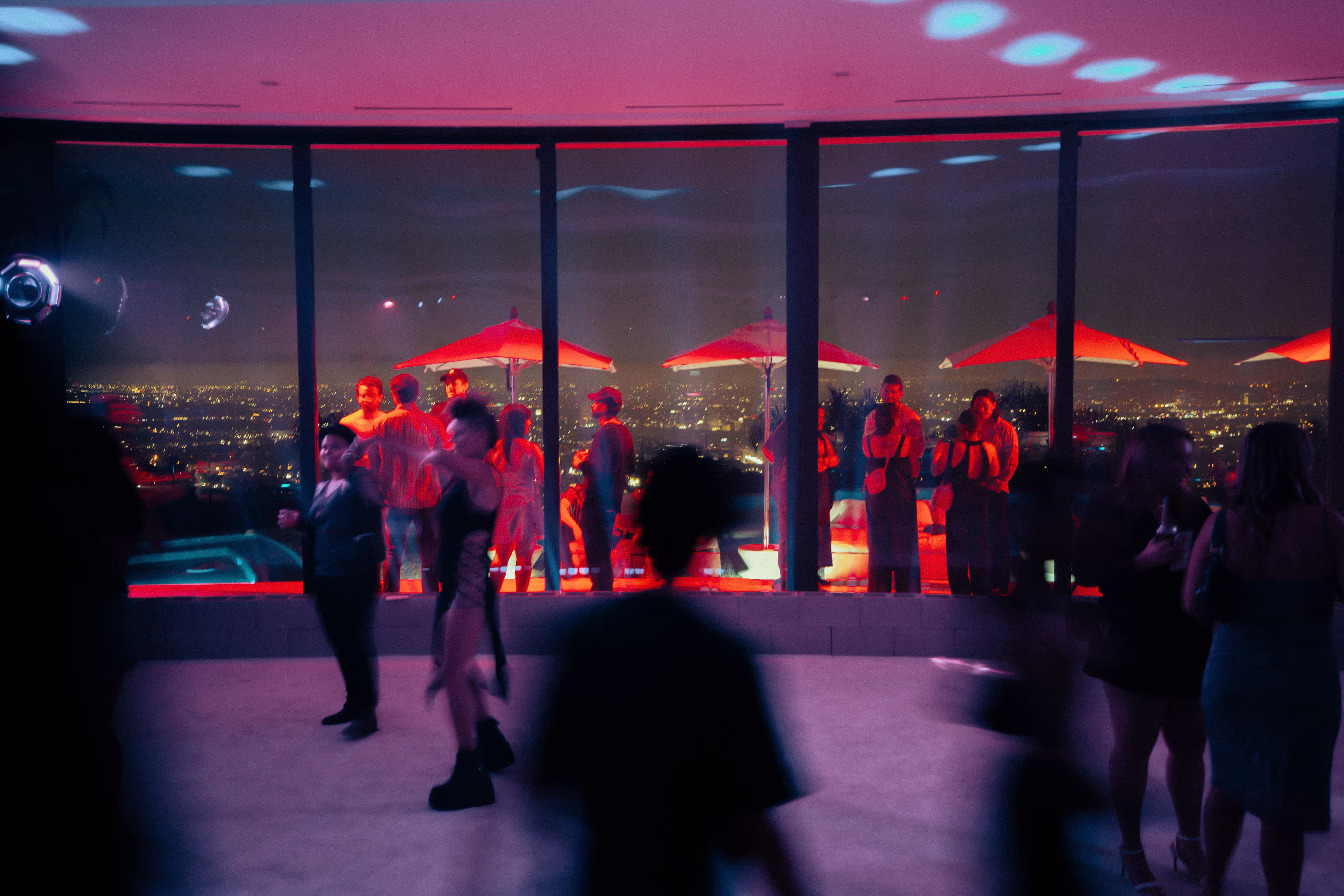 Nighttime rooftop party with people dancing and socializing against a city skyline backdrop lit by red and purple lights.