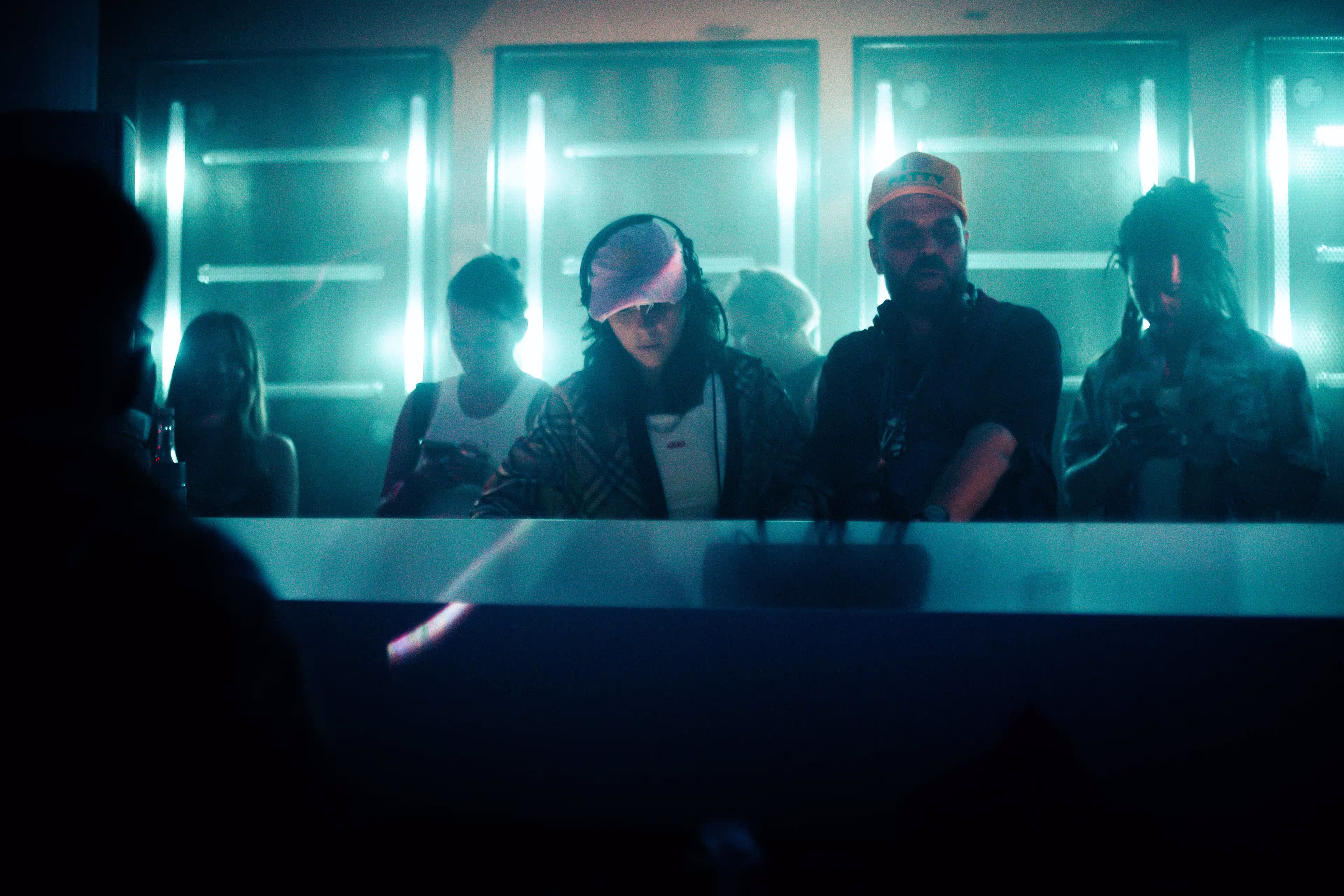 Group of people standing behind a DJ booth in a dimly lit club with blue lighting.