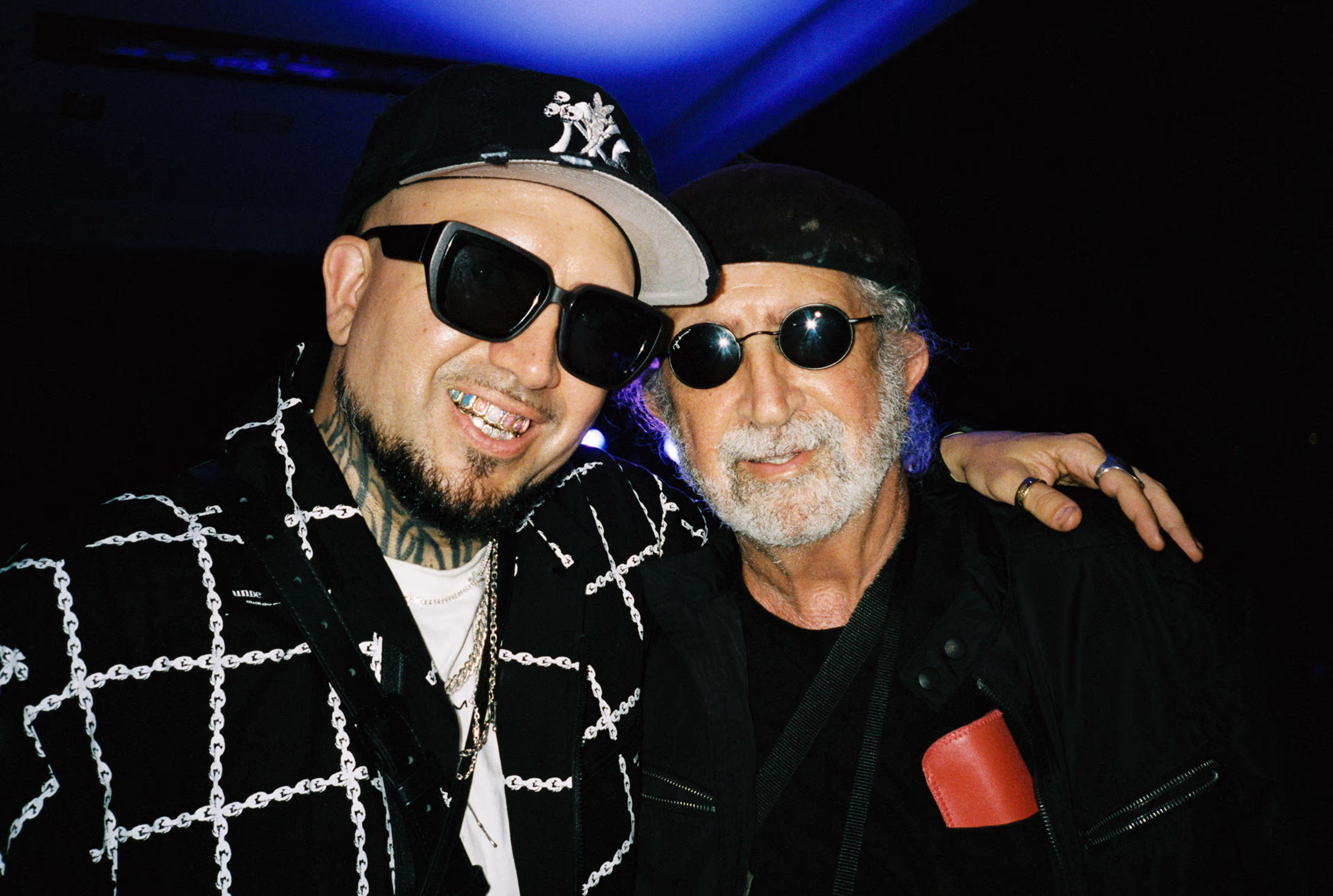 Two men posing closely together, both wearing sunglasses and black outfits.