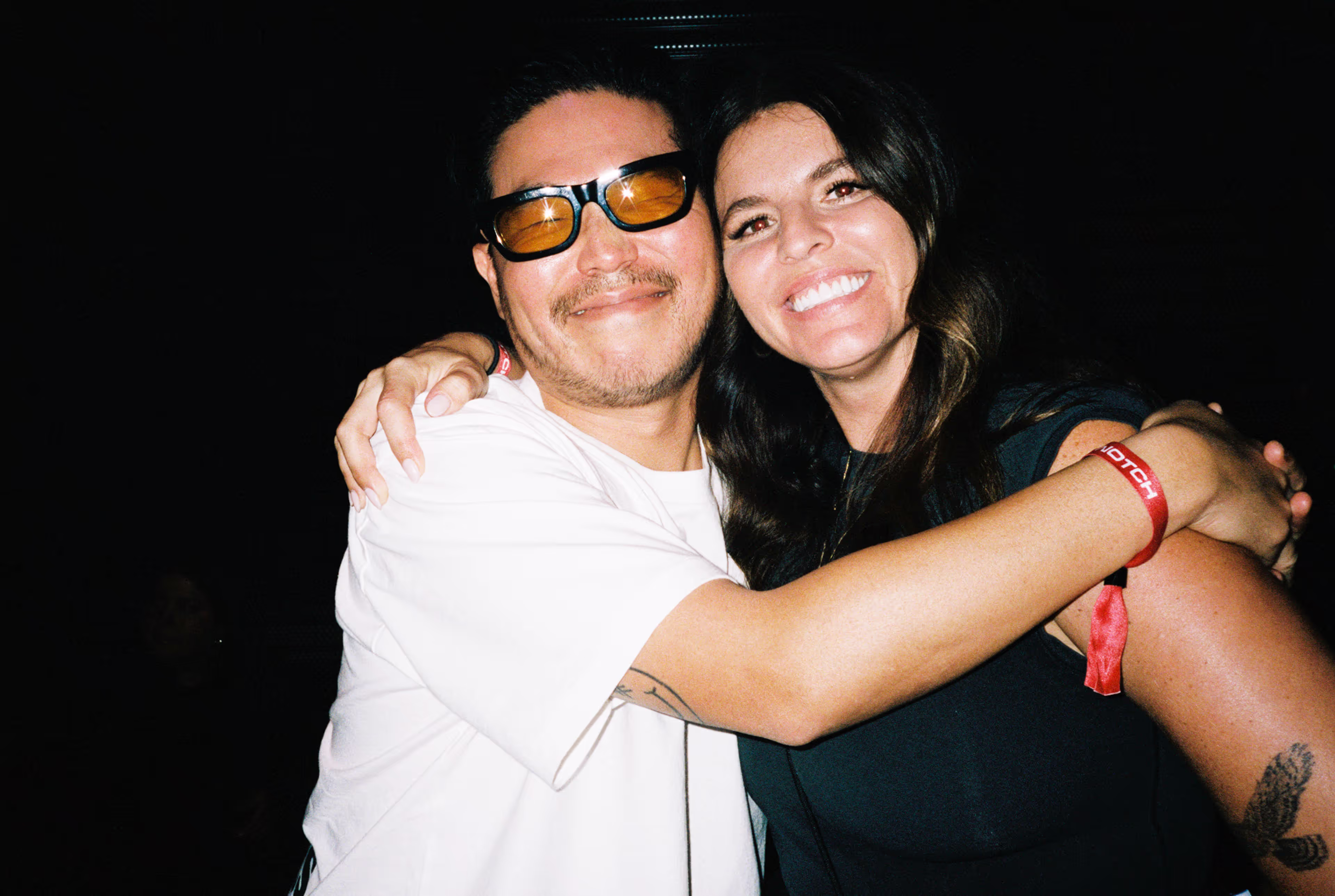Man wearing yellow sunglasses and white shirt hugs smiling woman with long dark hair.