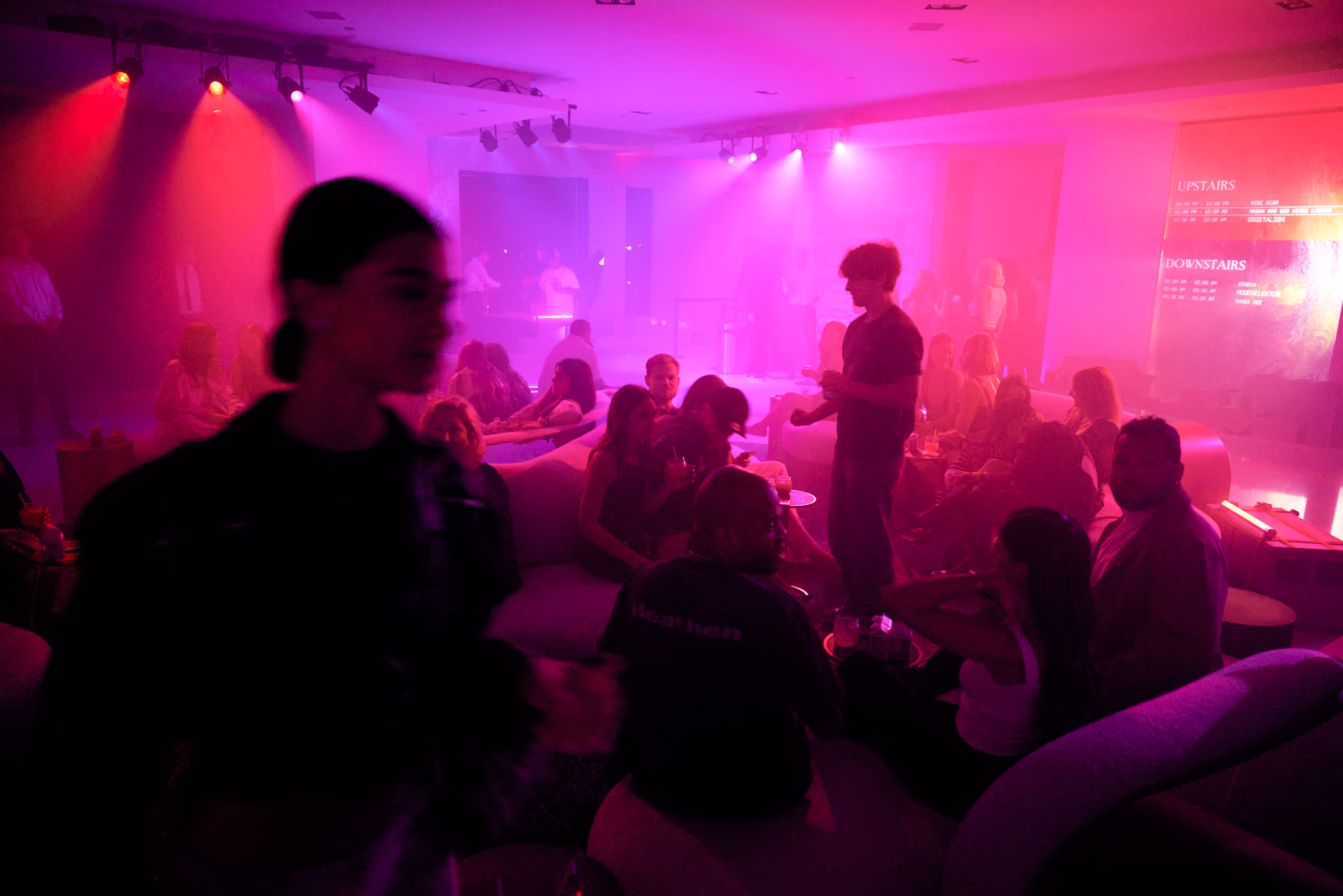 Dimly lit nightclub scene with groups of people sitting on couches and standing, illuminated by pink and purple lights.