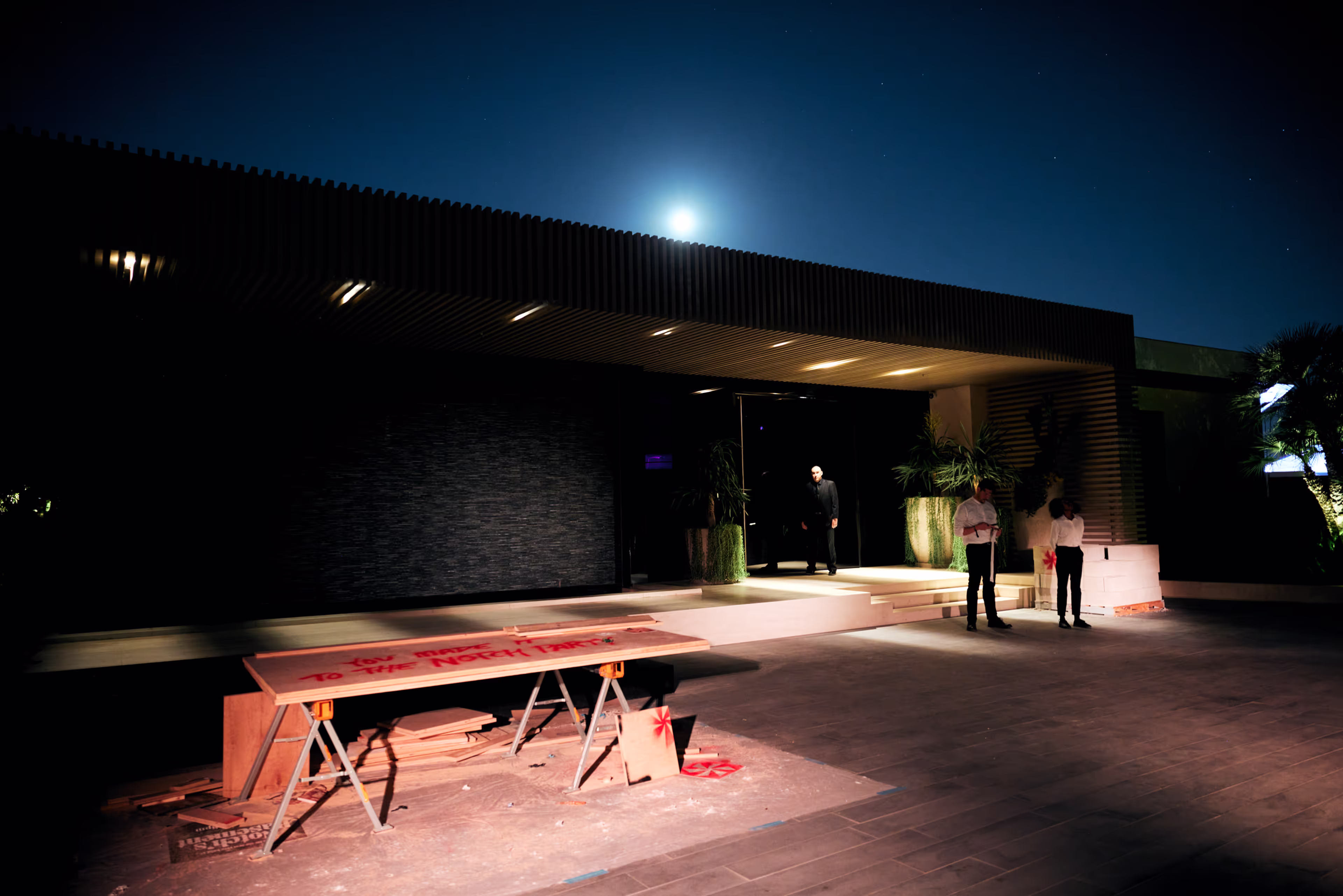 Nighttime scene of a modern building entrance with a security guard standing in the doorway and two people dressed in white shirts nearby, illuminated by overhead lights and moonlight.