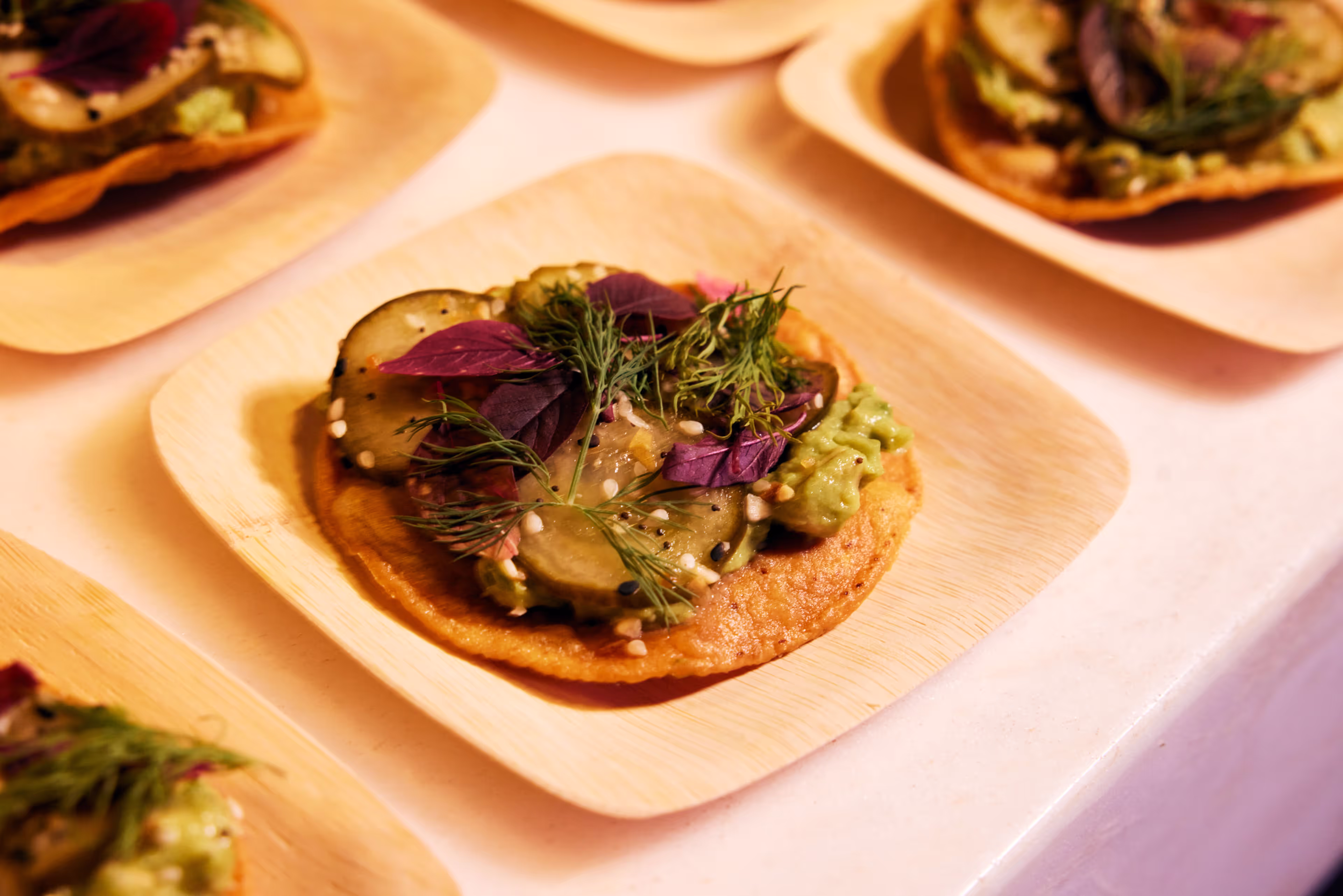 Personal-sized tostada topped with guacamole, sliced pickles, purple leaves, dill sprigs, and sesame seeds on a wooden plate.