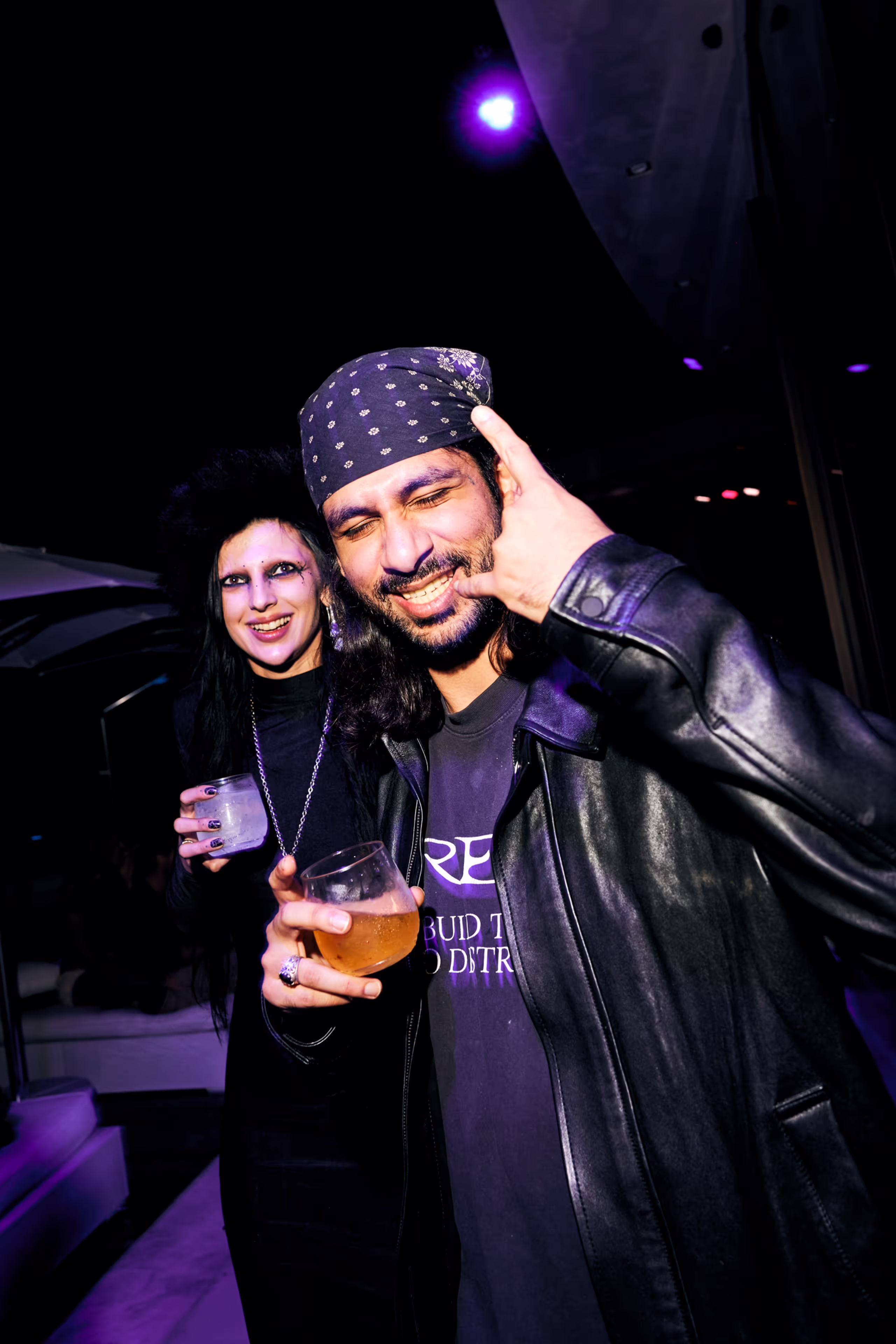 Man in a black leather jacket and bandana making a rock hand gesture and holding a drink, with a woman behind him in dark gothic makeup and clothing holding a glass.