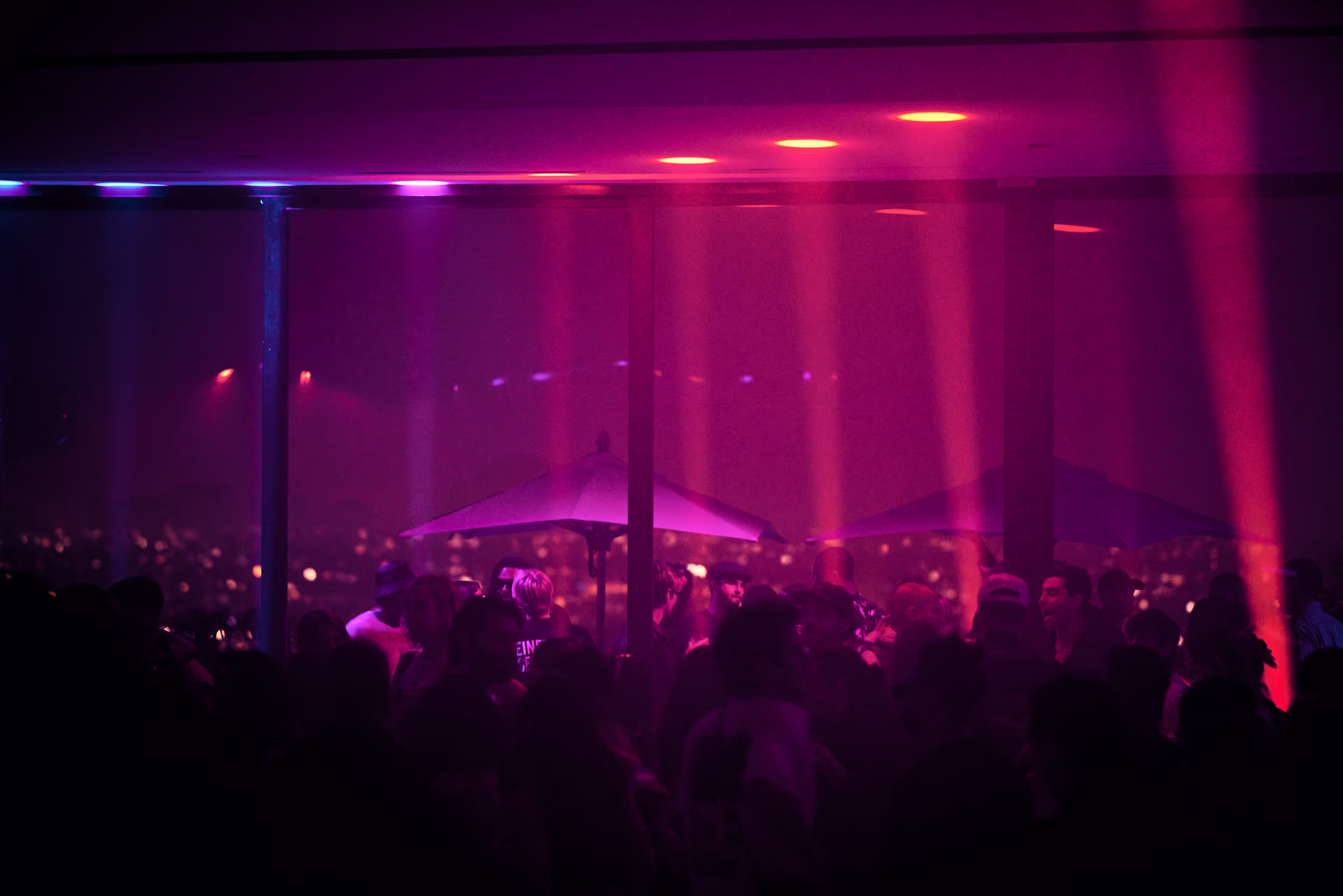 Crowded indoor party with purple and pink lighting and city lights visible through large windows.