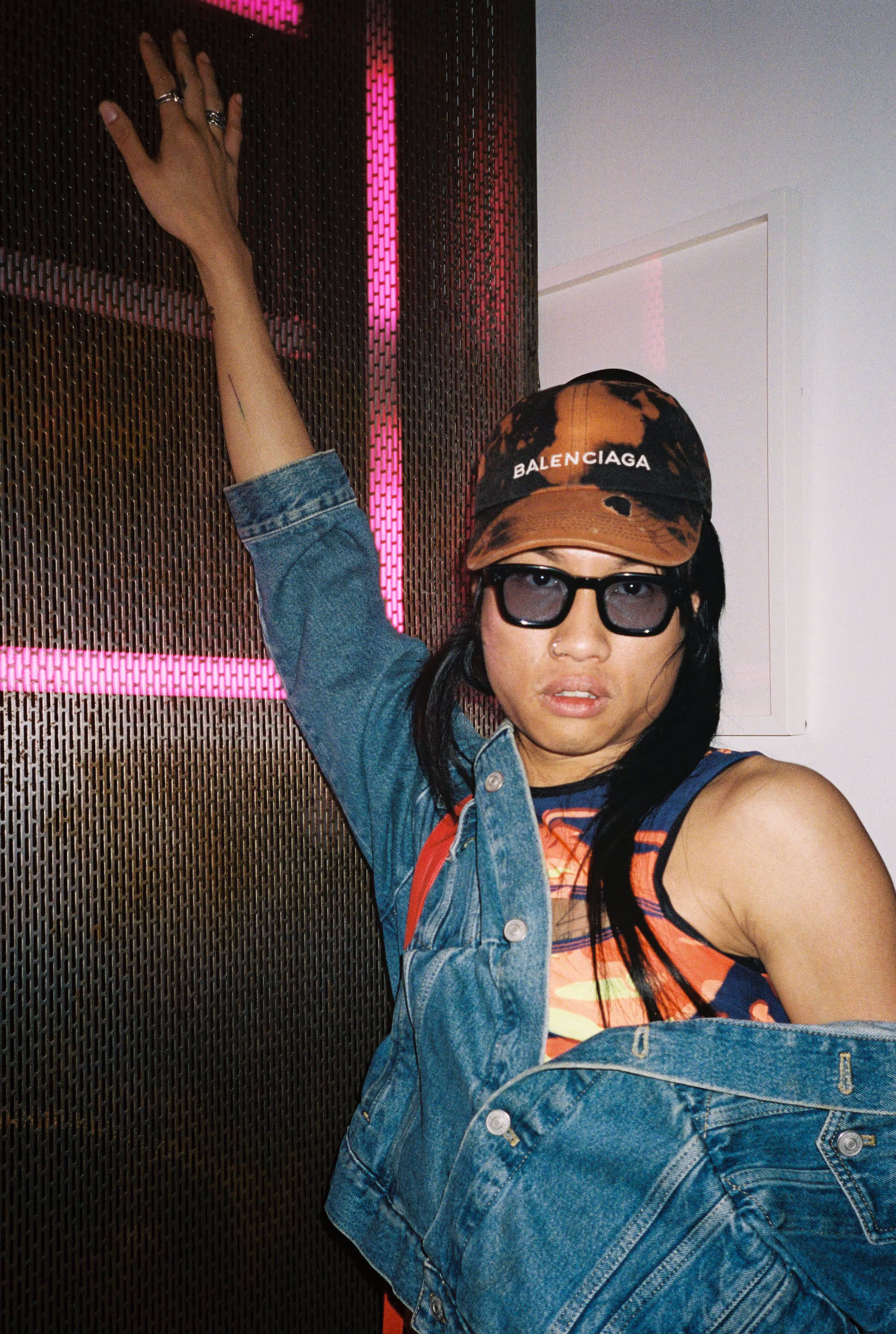 Person wearing a Balenciaga cap, sunglasses, colorful top, and denim jacket posing with one arm raised against a patterned wall with pink neon lights.