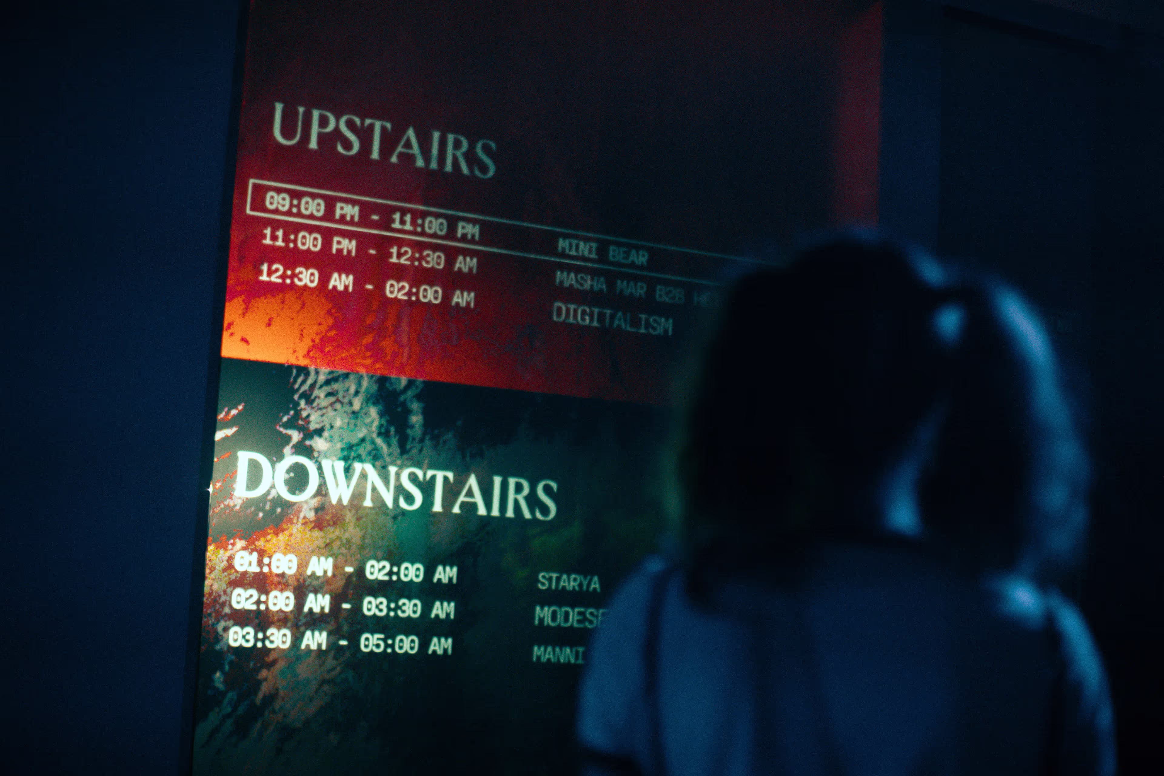 Person looking at a dimly lit schedule board listing upstairs and downstairs event times and performers.