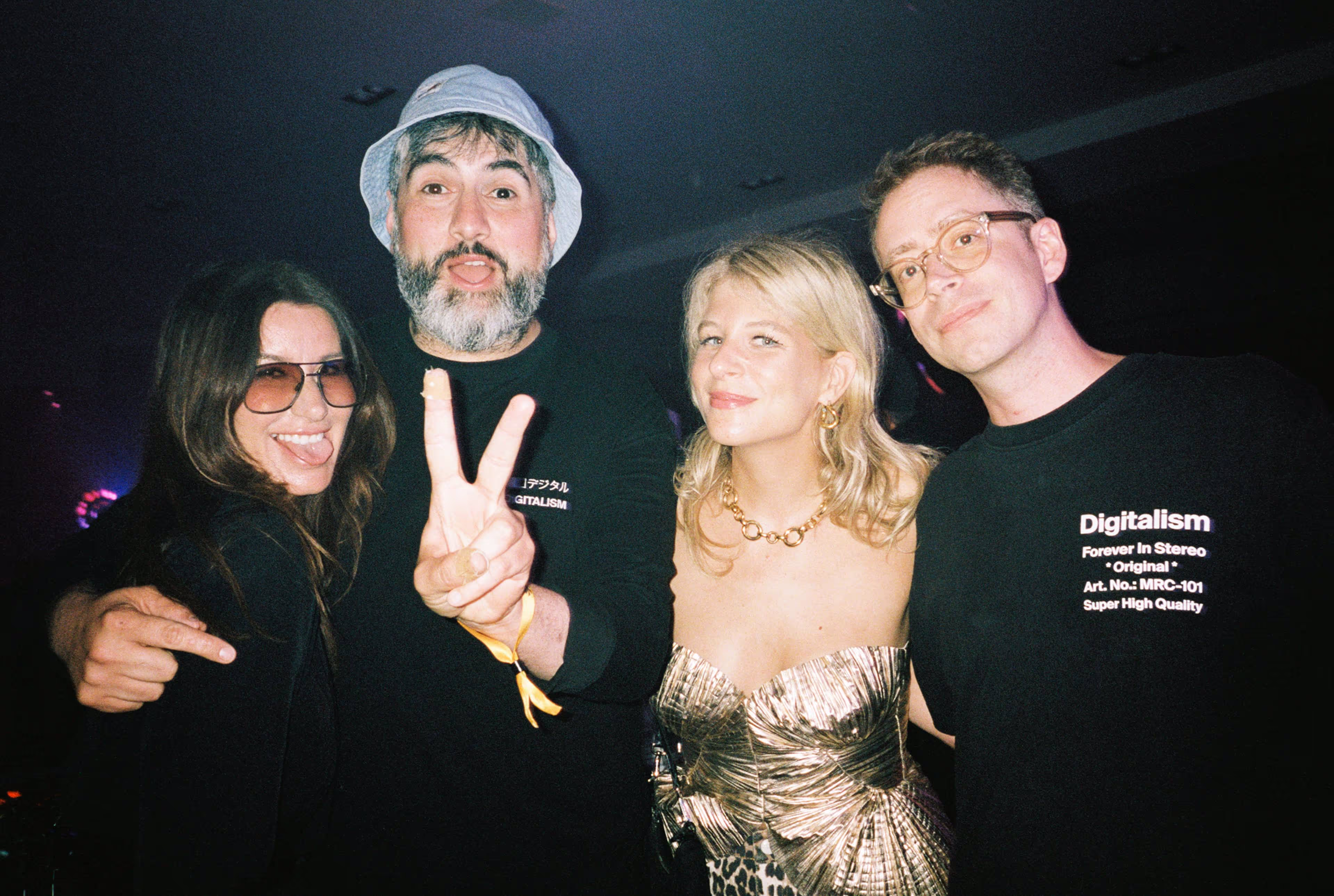 Four people posing at a nighttime event, one man wearing a bucket hat making a peace sign, a woman sticking out her tongue wearing sunglasses, another woman in a gold strapless dress, and a man in glasses wearing a Digitalism T-shirt.