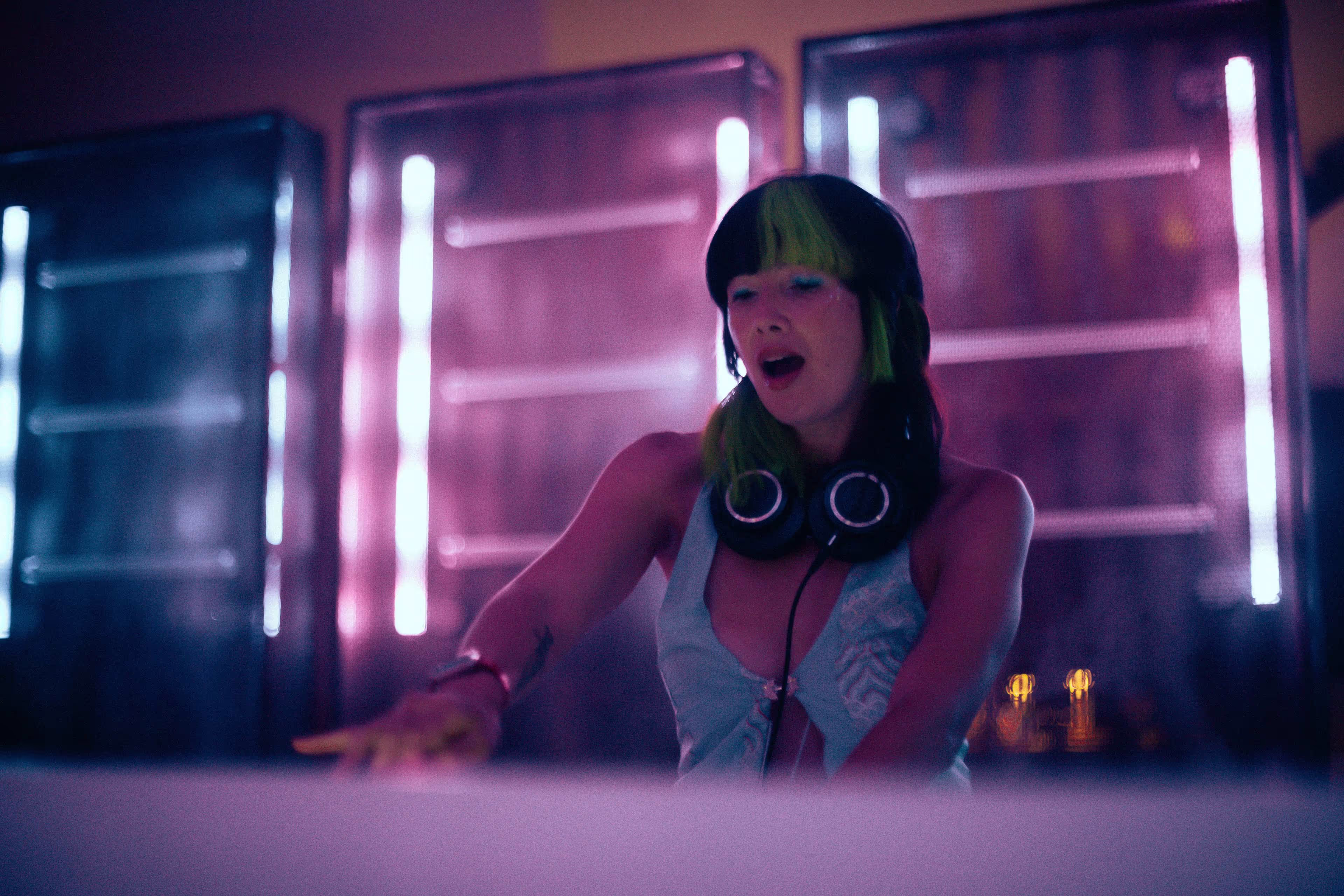 Female DJ with black and green hair wearing headphones and a light blue top, performing in a dimly lit club with purple and blue neon lights.