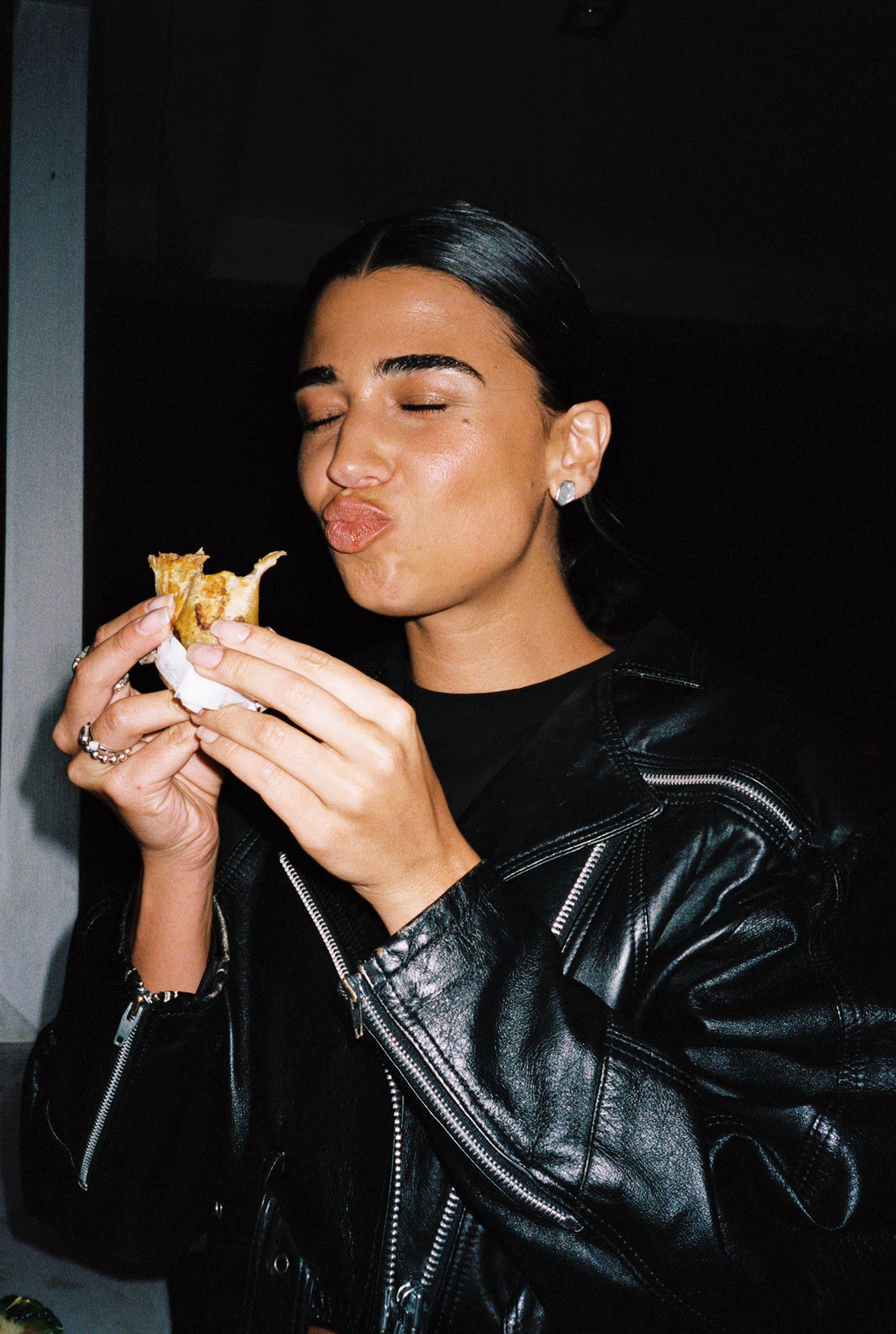 Person with slicked-back hair and black leather jacket savoring a bite of food with eyes closed.
