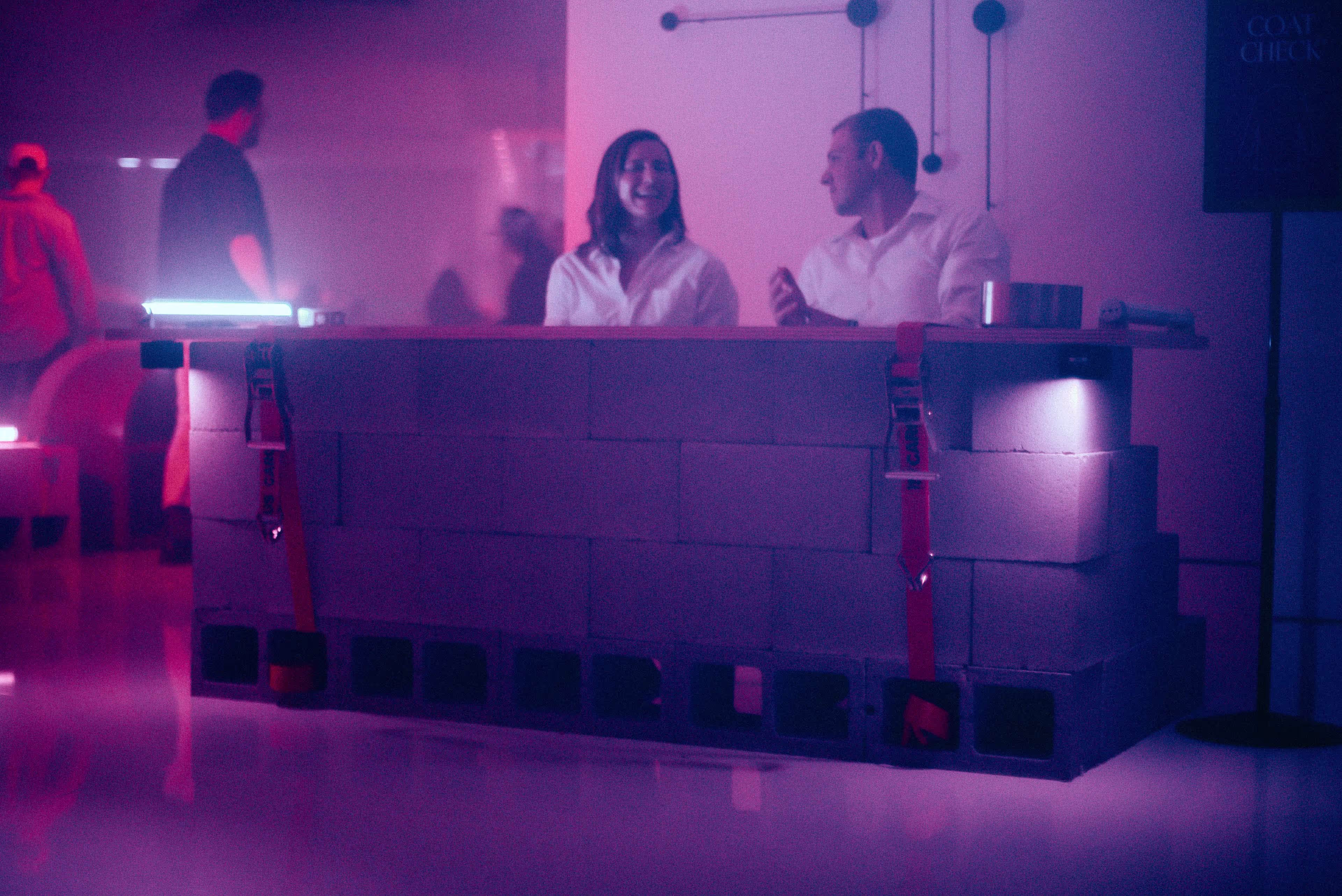 Two people in white shirts standing behind a bar made of stacked cinder blocks with red straps in a dimly lit purple room.