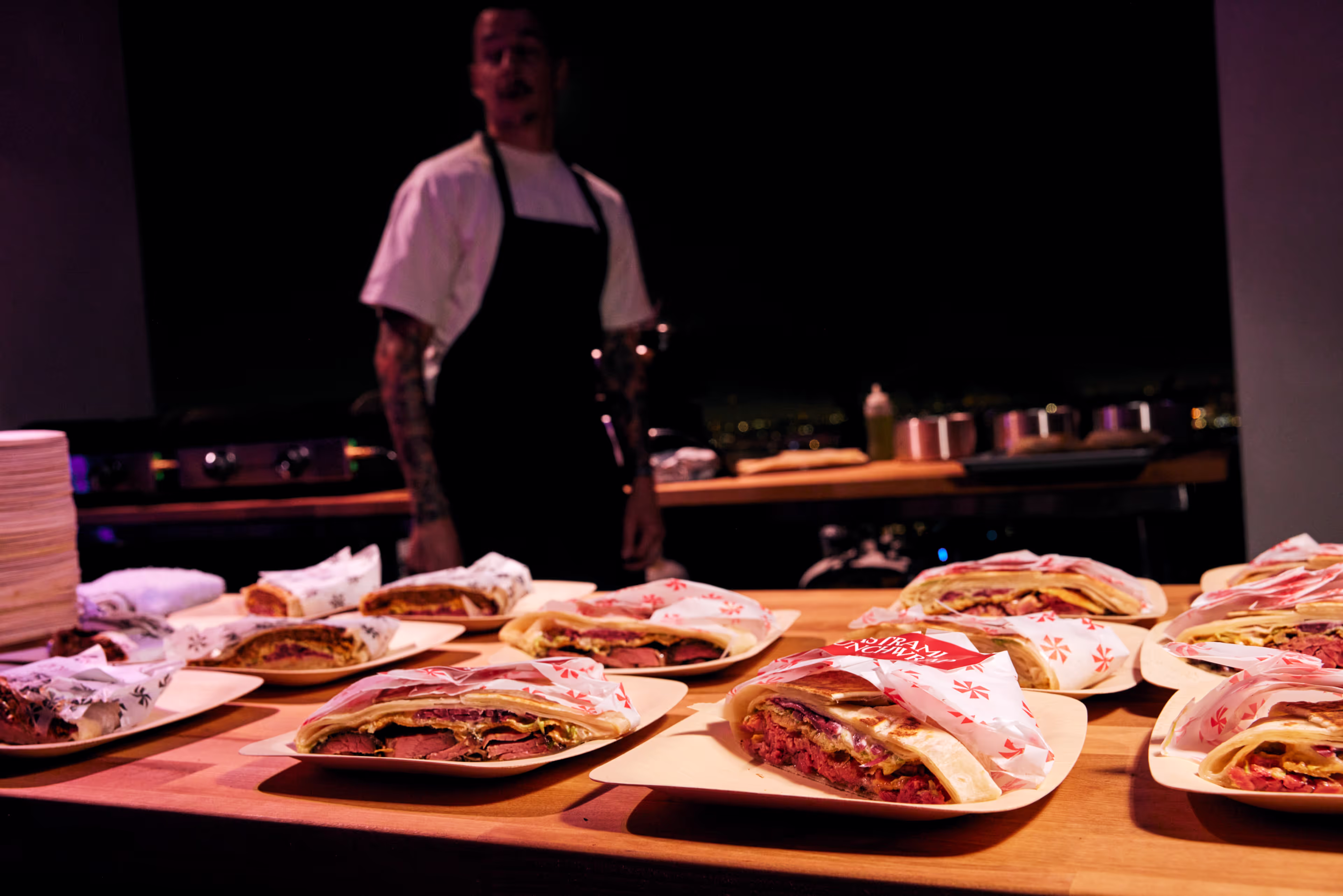 Assorted sandwiches wrapped in branded paper on plates on a wooden counter with a chef in a black apron standing in the background.