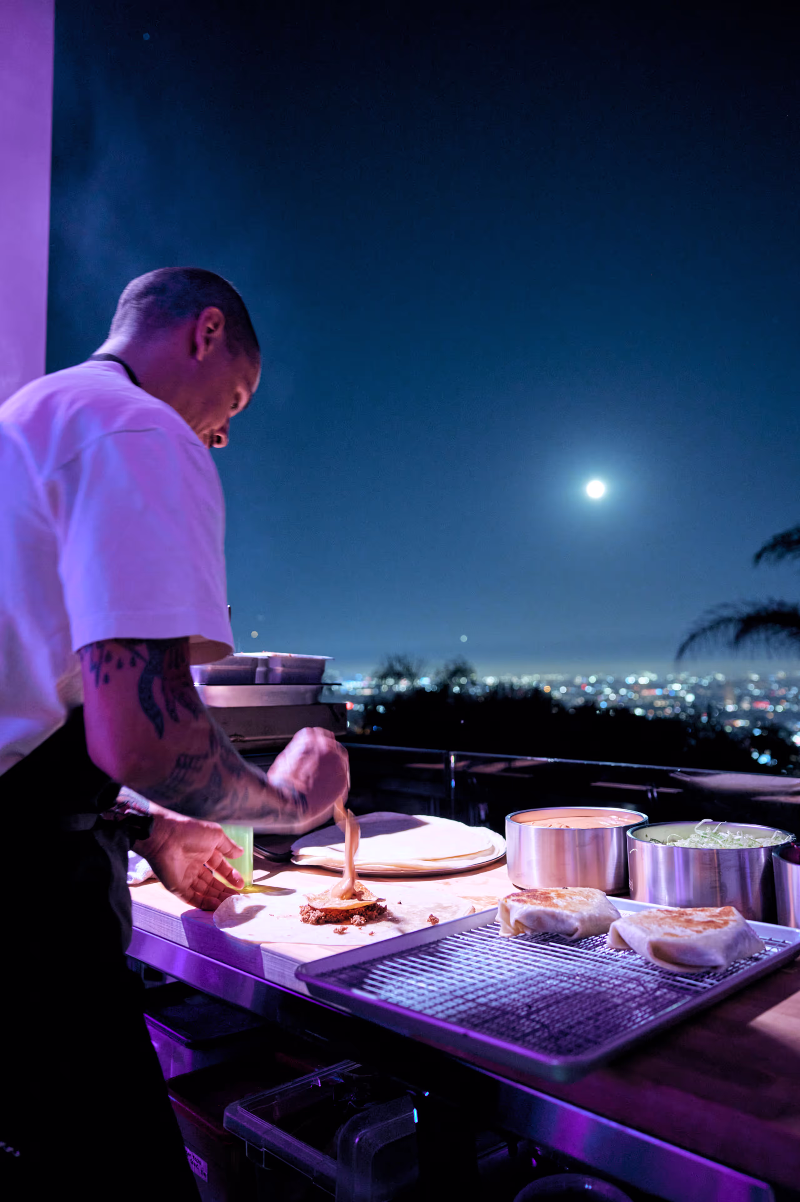 Chef preparing food on a countertop at night with city lights and a full moon in the background.