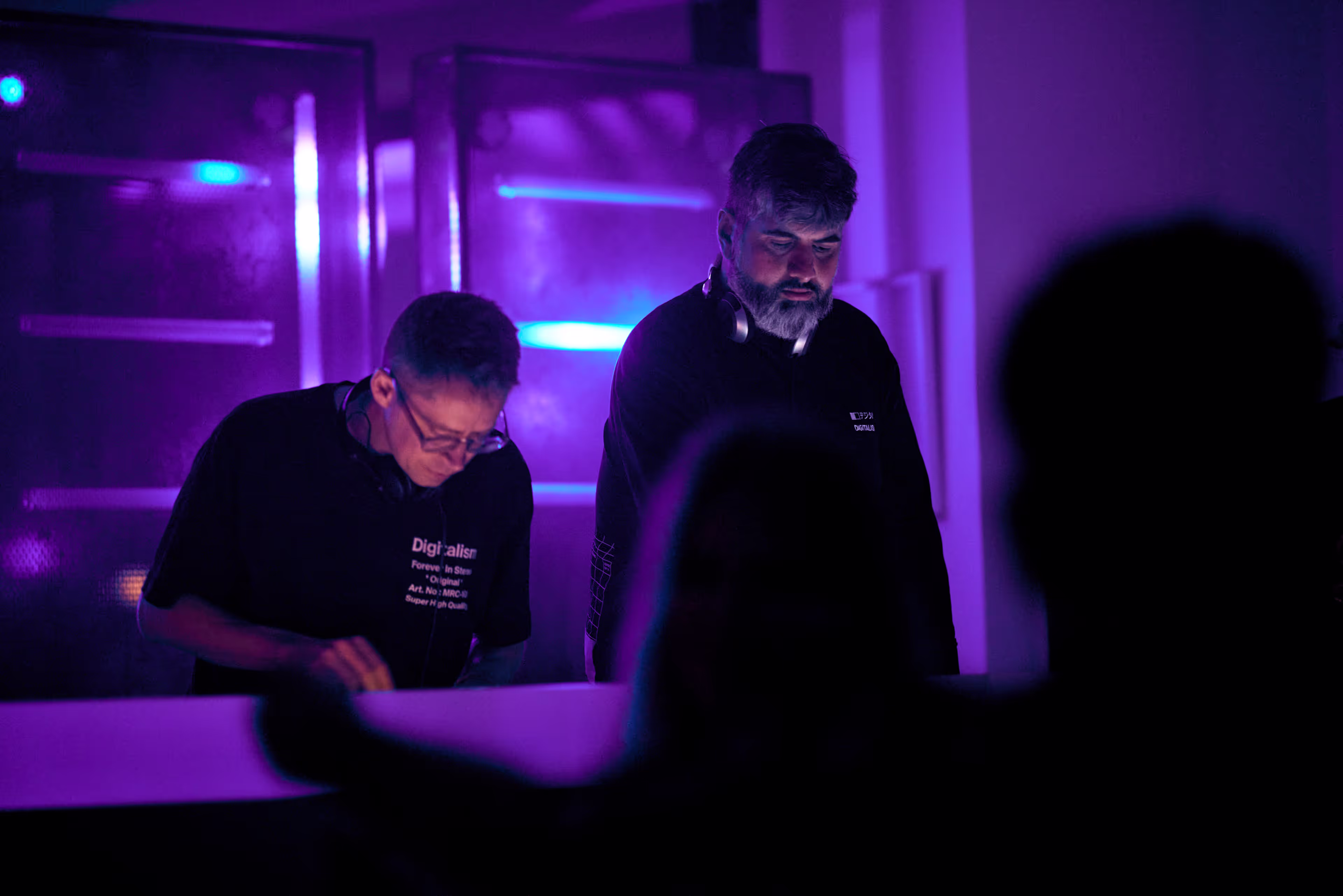 Two DJs performing under purple and blue lighting with silhouettes of audience members in the foreground.