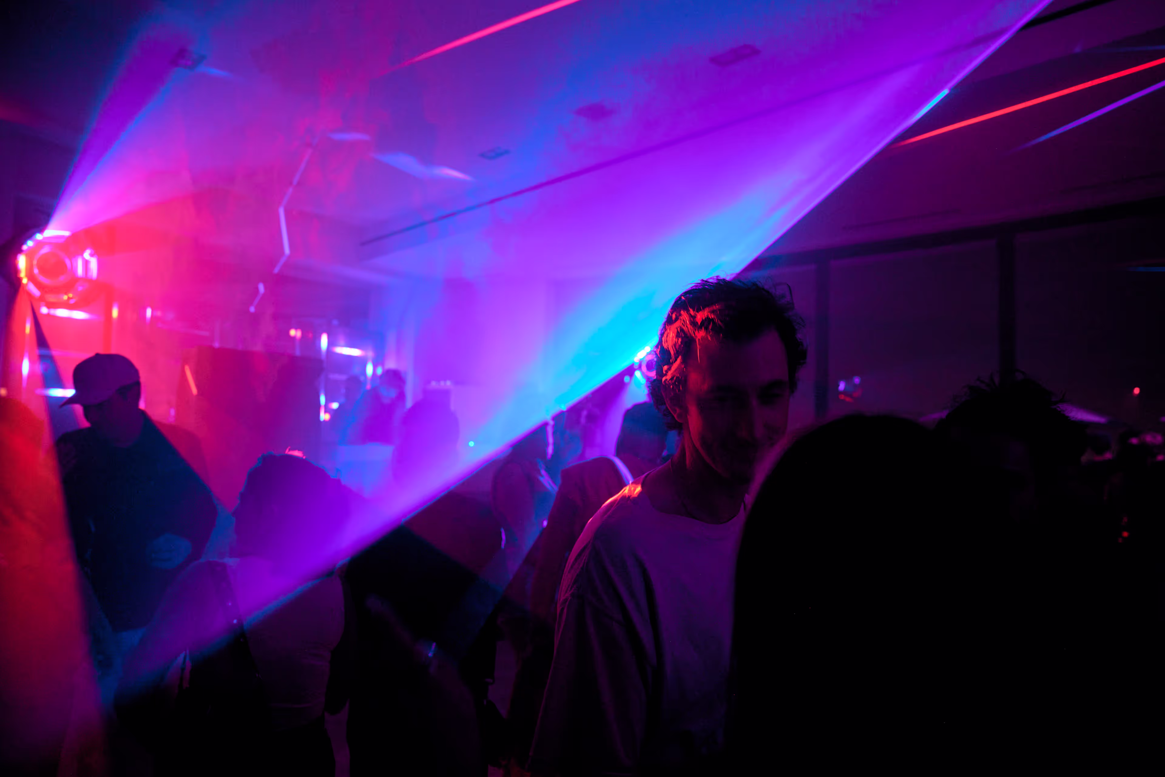 Dimly lit nightclub scene with people dancing and colorful pink and blue laser lights cutting through smoke.