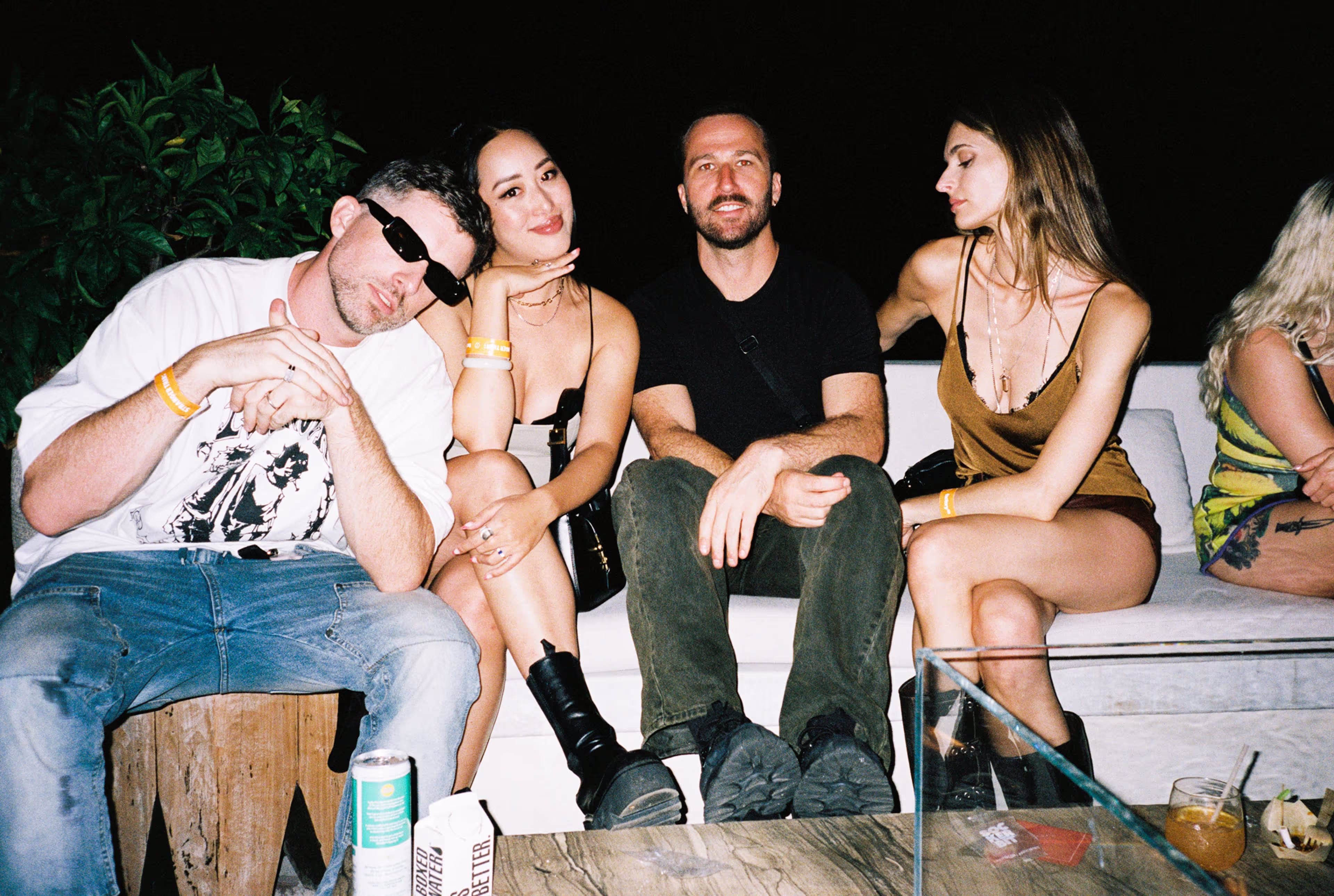 Four people sitting on a white couch at night with drinks on a table.
