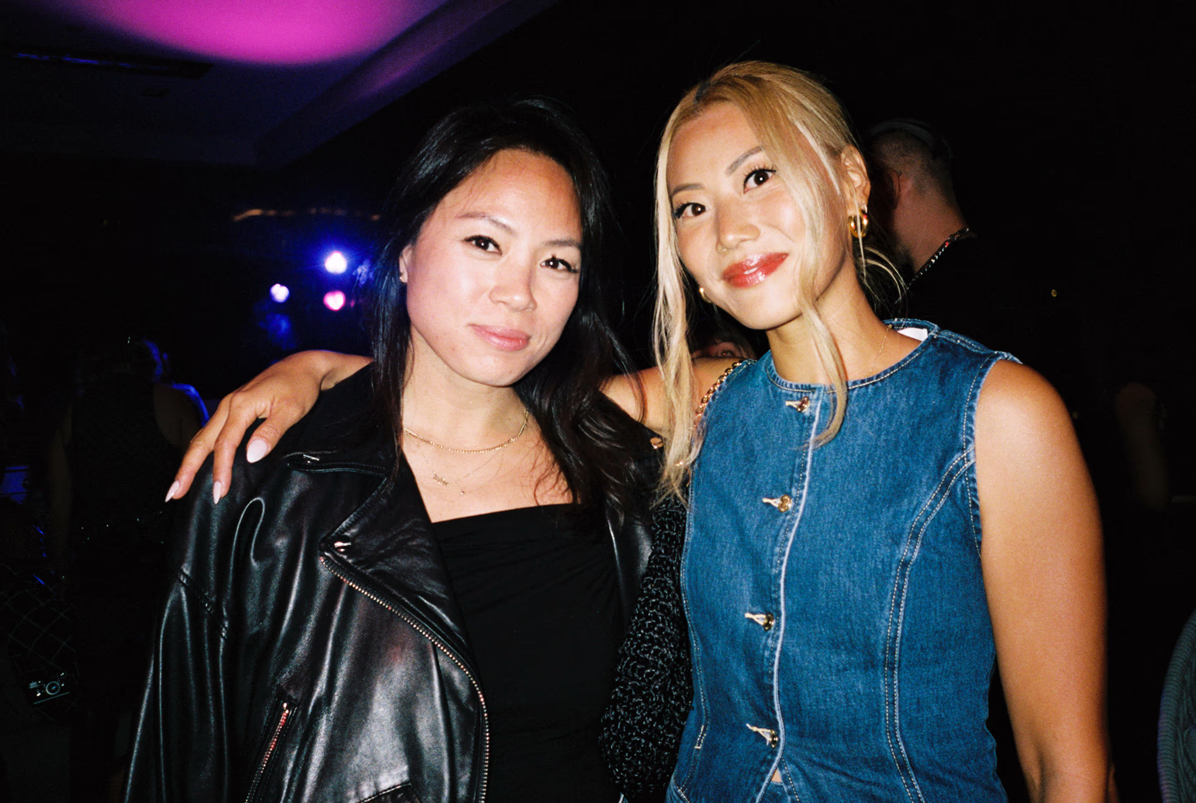 Two women smiling, one in a black leather jacket and the other in a sleeveless denim dress.