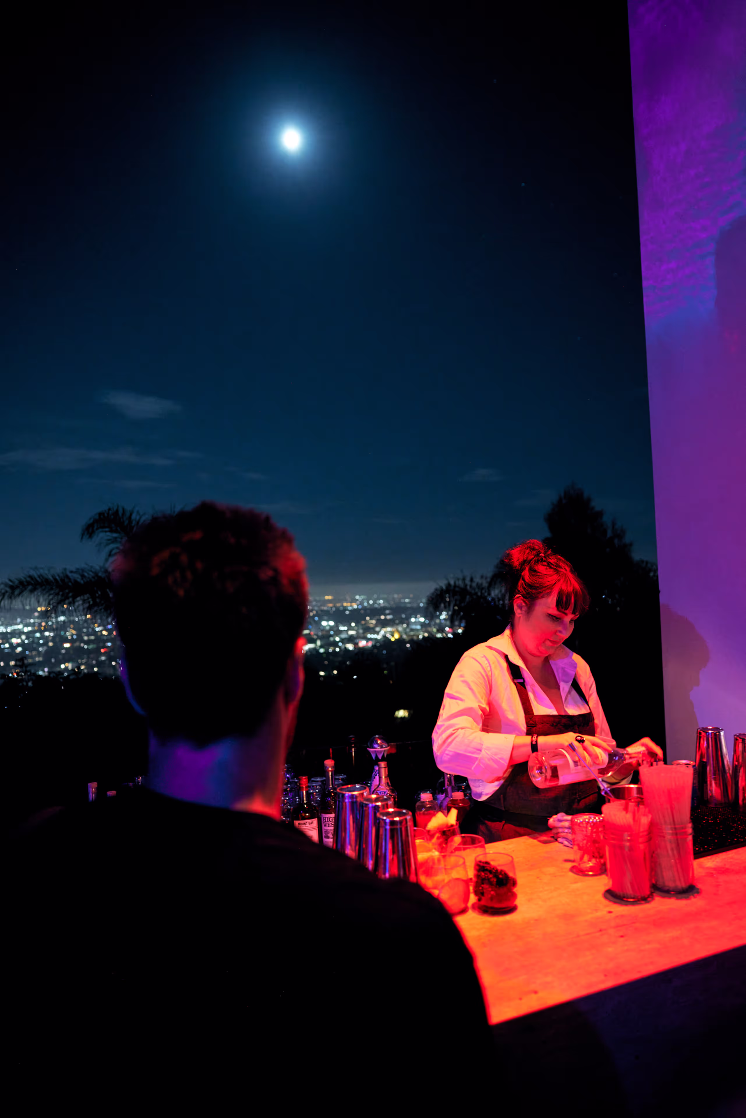 Bartender pouring a drink at an outdoor bar at night with a city skyline and full moon in the background.