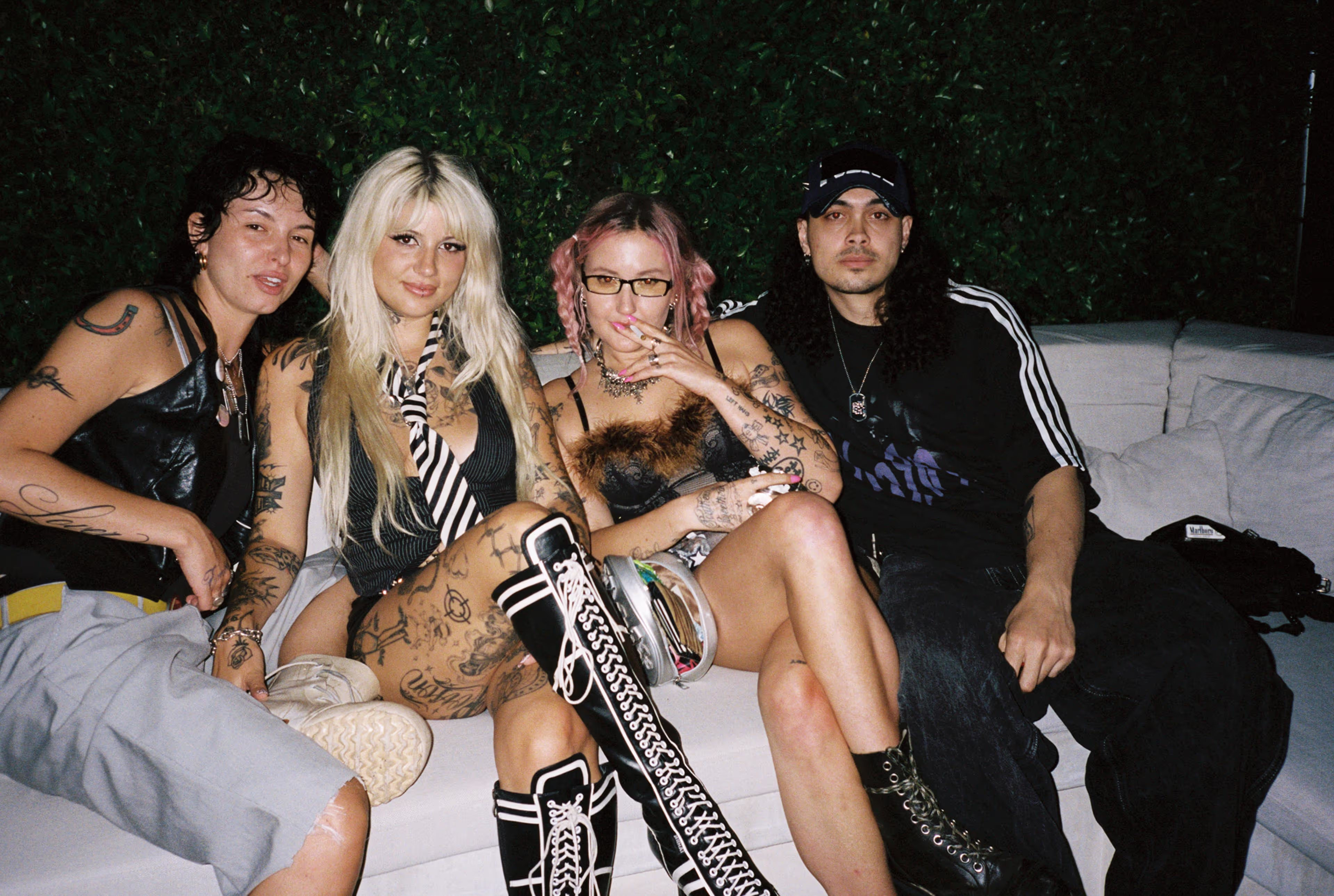 Four tattooed friends sitting closely on a white couch at night with a green hedge background.